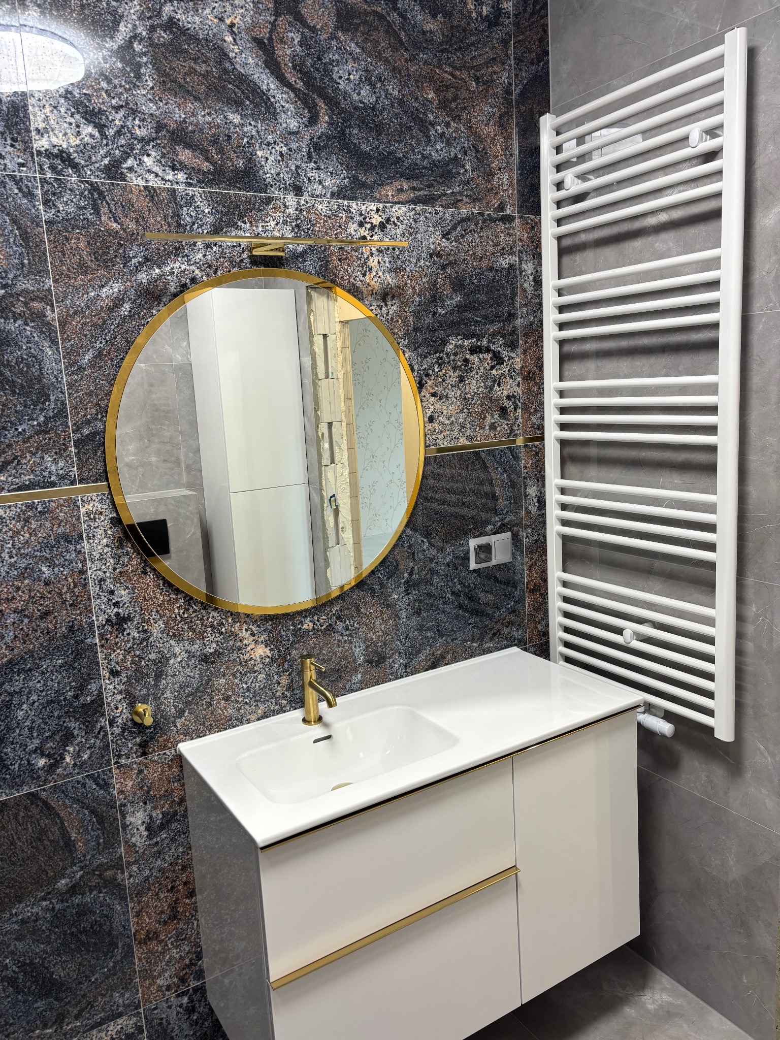 Wykończona łazienka: ciemna, wzorzysta ściana z lustrem w złotej ramie, biała szafka z umywalką i złota bateria, biały grzejnik drabinkowy na szarej ścianie.