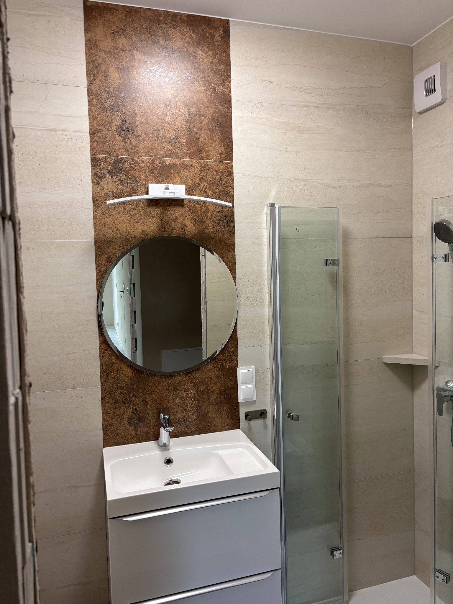 Nowoczesna łazienka po remoncie: biała szafka z umywalką, okrągłe lustro z oświetleniem, kabina prysznicowa z chromowanymi detalami i beżowe płytki imitujące kamień.