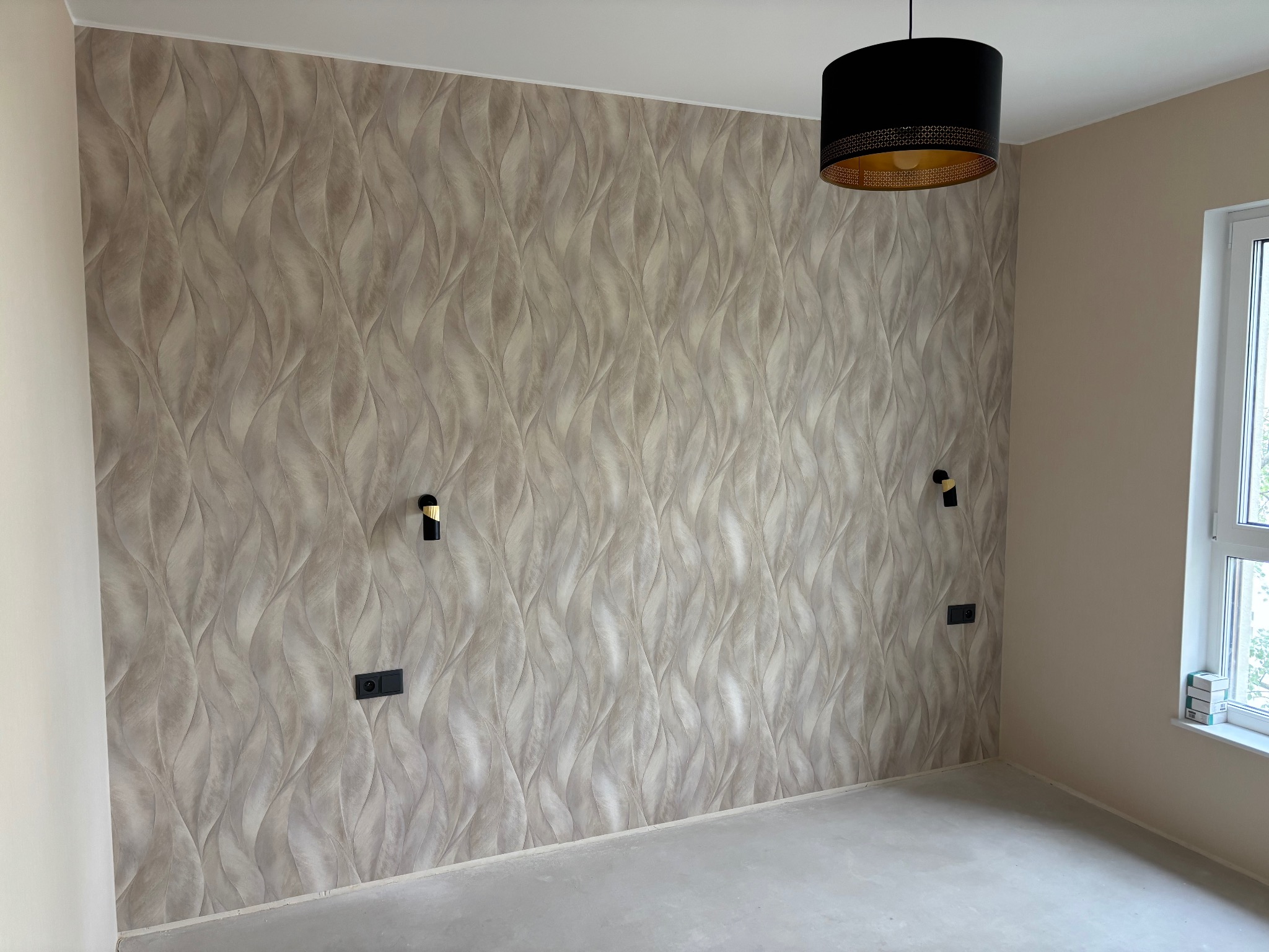 Wykończone wnętrze pokoju z tapetą w liście, czarnymi gniazdkami i lampą. Ściany pomalowane na beżowo, surowa podłoga. Widoczne okno.