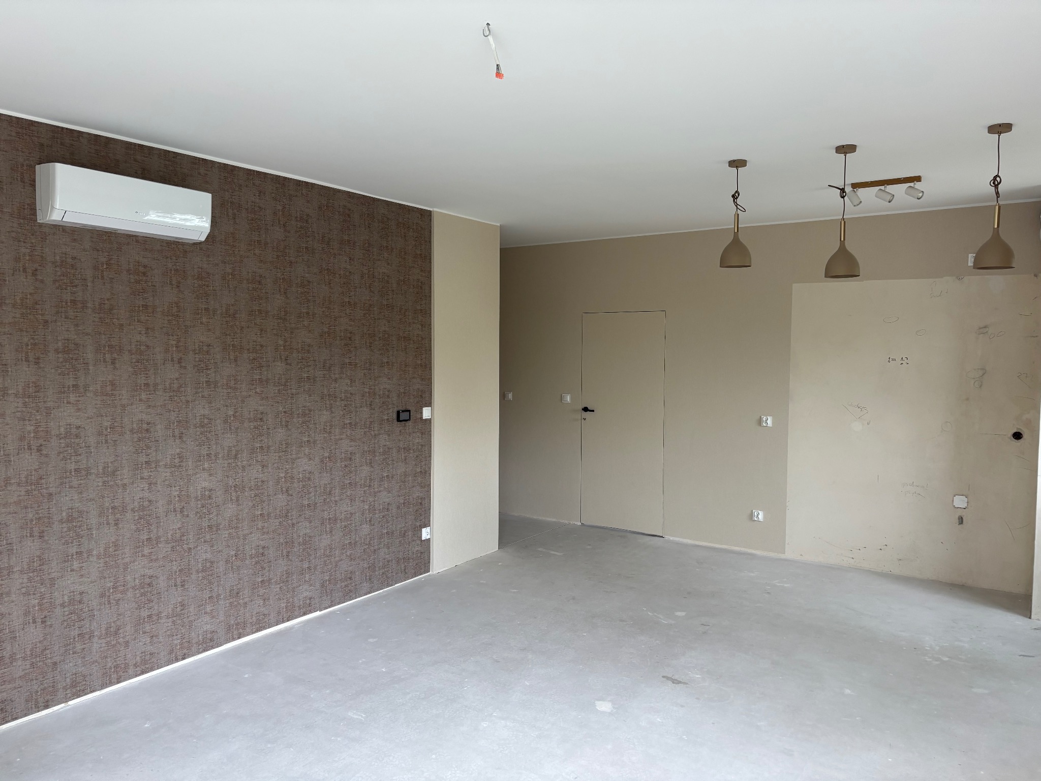 Wnętrze pokoju po remoncie z klimatyzatorem na ścianie z brązową tapetą. Widoczne lampy sufitowe, gładzie i wylewka podłogowa. Neutralna kolorystyka ścian.