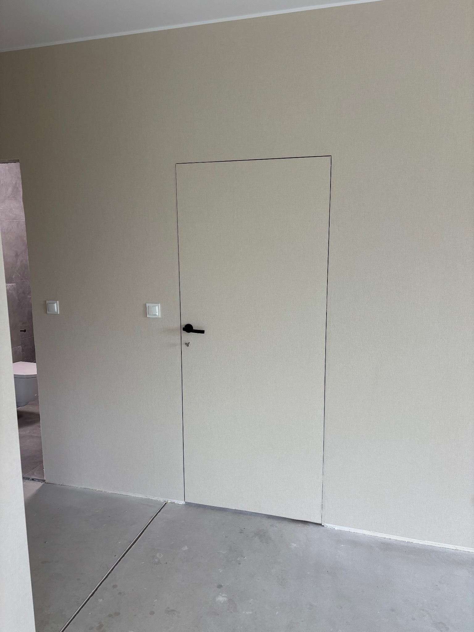 Minimalistyczne wnętrze: gładkie, beżowe ściany, ukryte drzwi z czarną klamką, widoczny fragment łazienki z białą ceramiką. Surowy beton na podłodze.