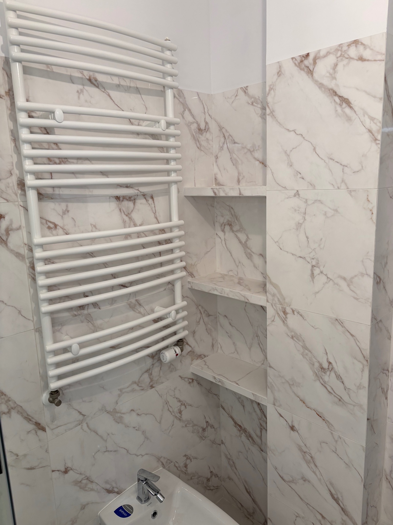 Biały grzejnik drabinkowy na ścianie z marmurowymi płytkami w łazience. W tle wnęka z półkami, umywalka z baterią. Ujęcie z dołu.