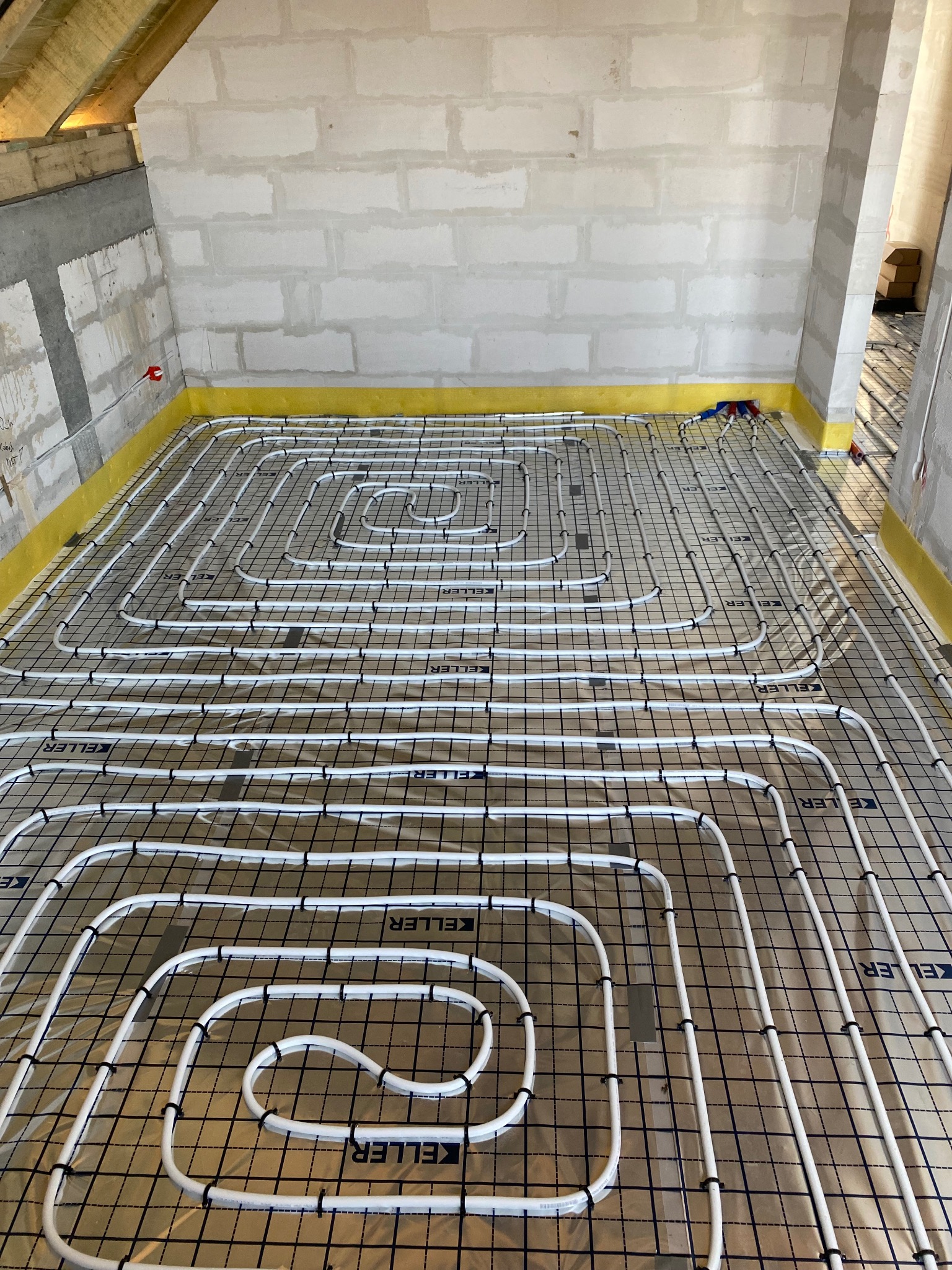 Białe rury ogrzewania podłogowego marki Keller, ułożone na folii z czarną siatką, przytwierdzone spinkami, w surowym wnętrzu z żółtą taśmą przy ścianach.