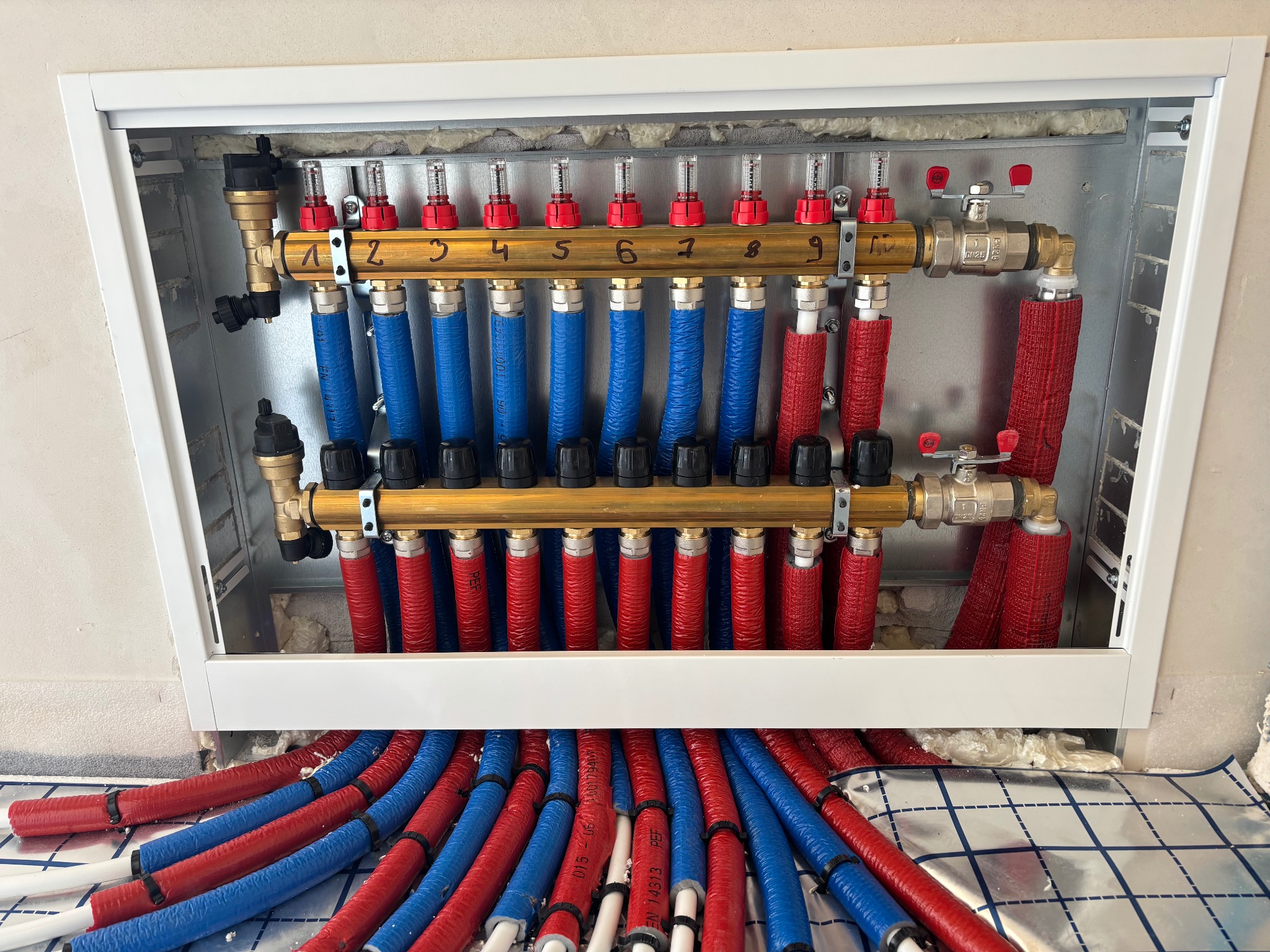Rozdzielacz ogrzewania podłogowego z numerowanymi zaworami, rurami w izolacji czerwonej i niebieskiej, zamontowany w białej szafce. Widoczna folia izolacyjna na podłodze.