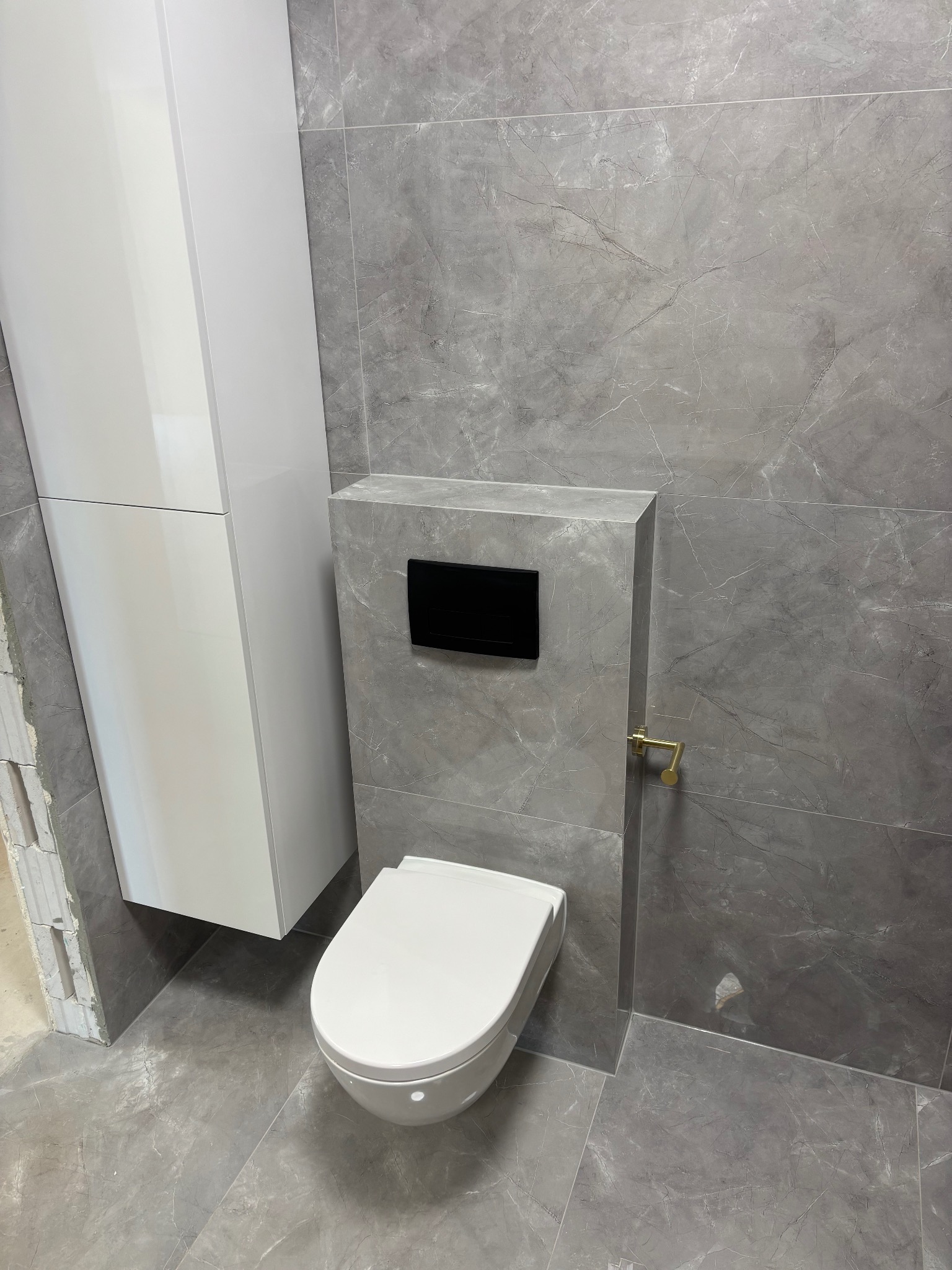 Nowoczesna toaleta podwieszana z szarą marmurową okładziną, czarnym przyciskiem i złotym uchwytem na papier, obok biała szafka w łazience po remoncie.