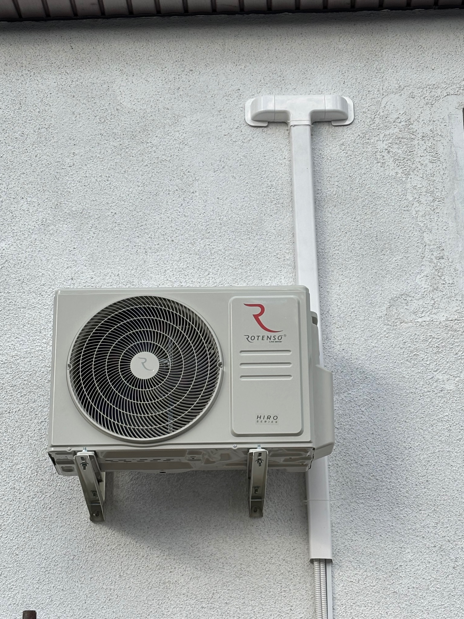 Zamontowana jednostka zewnętrzna klimatyzacji Rotenso Hiro Series na teksturowanej, jasnej ścianie budynku z widocznymi wspornikami i odprowadzeniem skroplin.