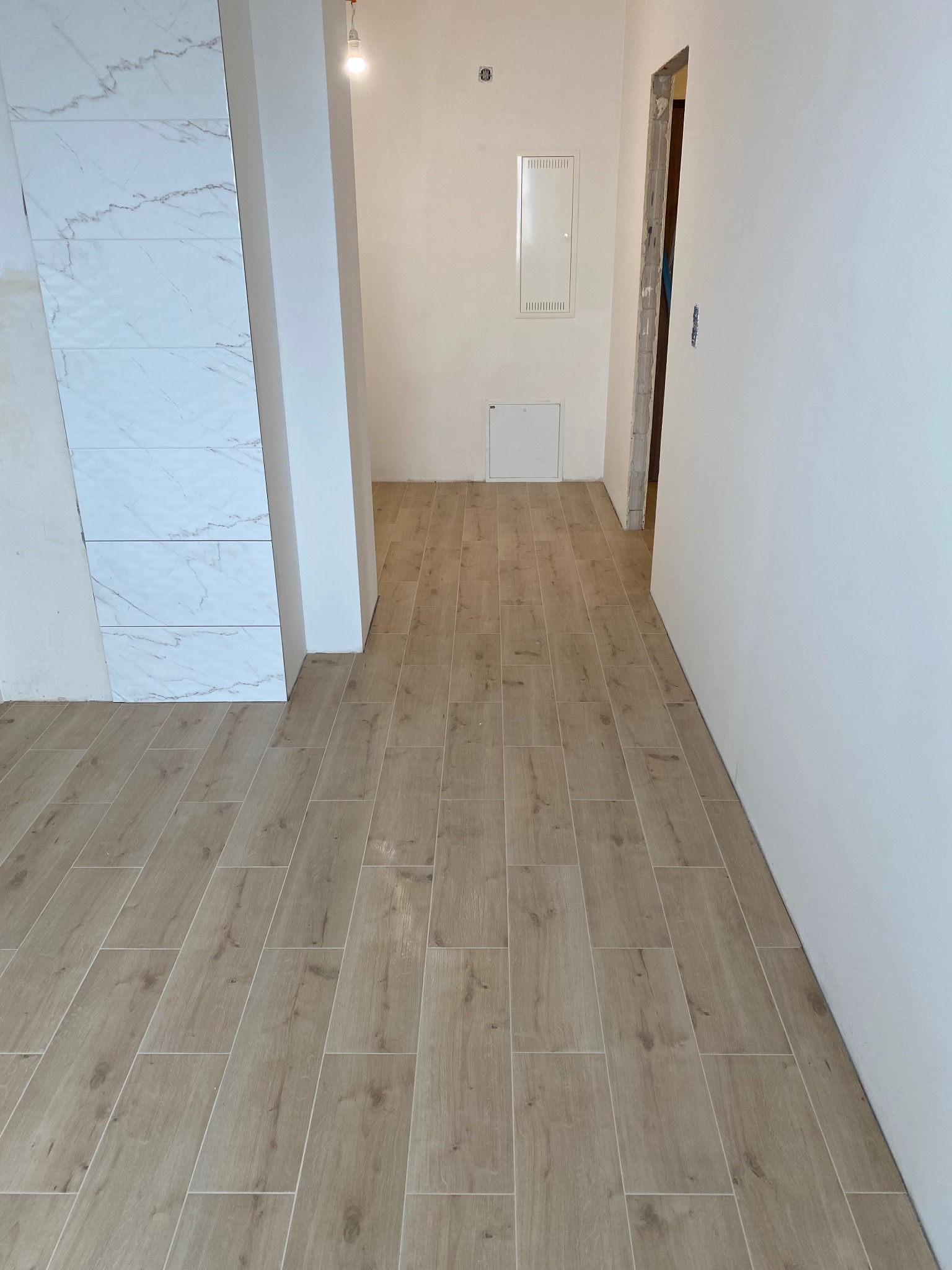 Ukończona podłoga z płytek imitujących drewno w jasnym odcieniu, kontrastująca z białą ścianą i fragmentem marmurowej okładziny. Widoczny fragment instalacji elektrycznej.