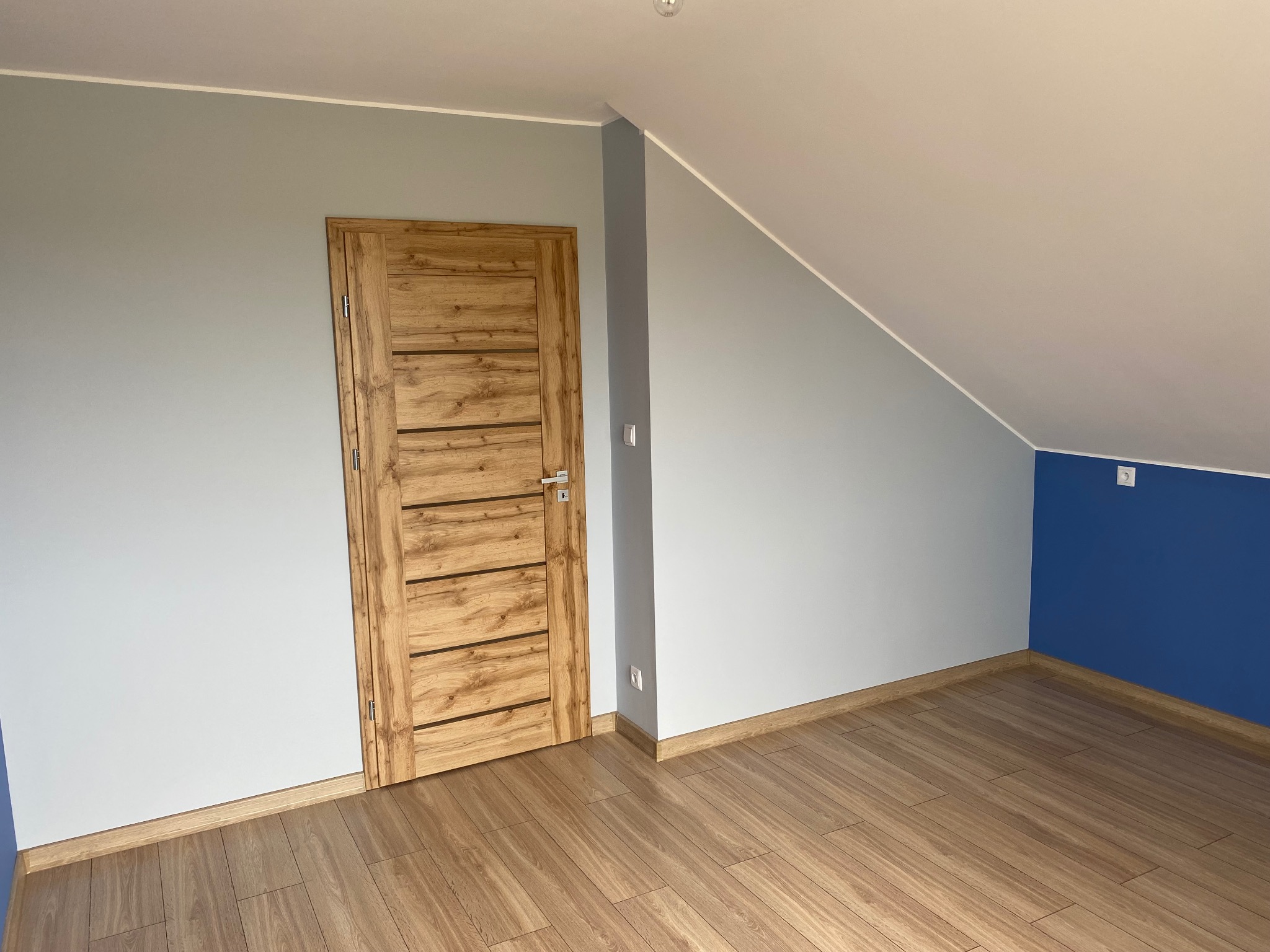 Wykończone wnętrze poddasza: drewniana podłoga i drzwi z poziomymi panelami, szare ściany z akcentem granatowym, biały sufit. Widok na fragment pokoju.