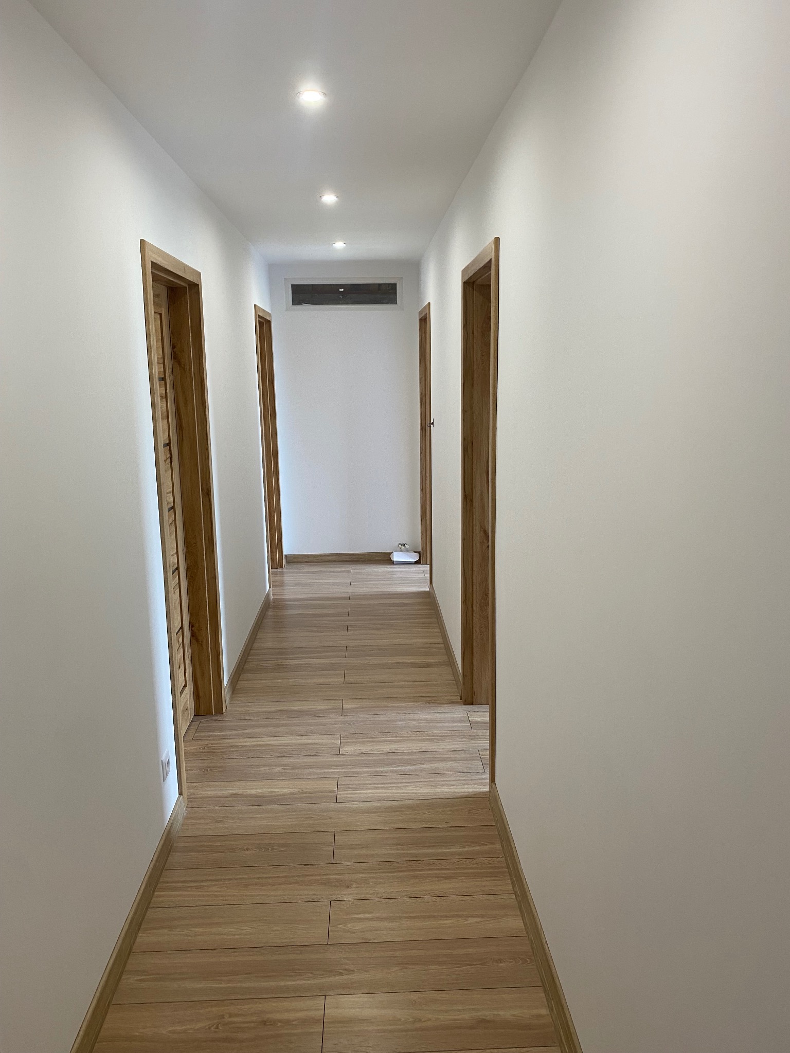 Jasny korytarz z panelami podłogowymi imitującymi drewno. Białe ściany, drewniane futryny drzwi. Widoczne oświetlenie sufitowe.