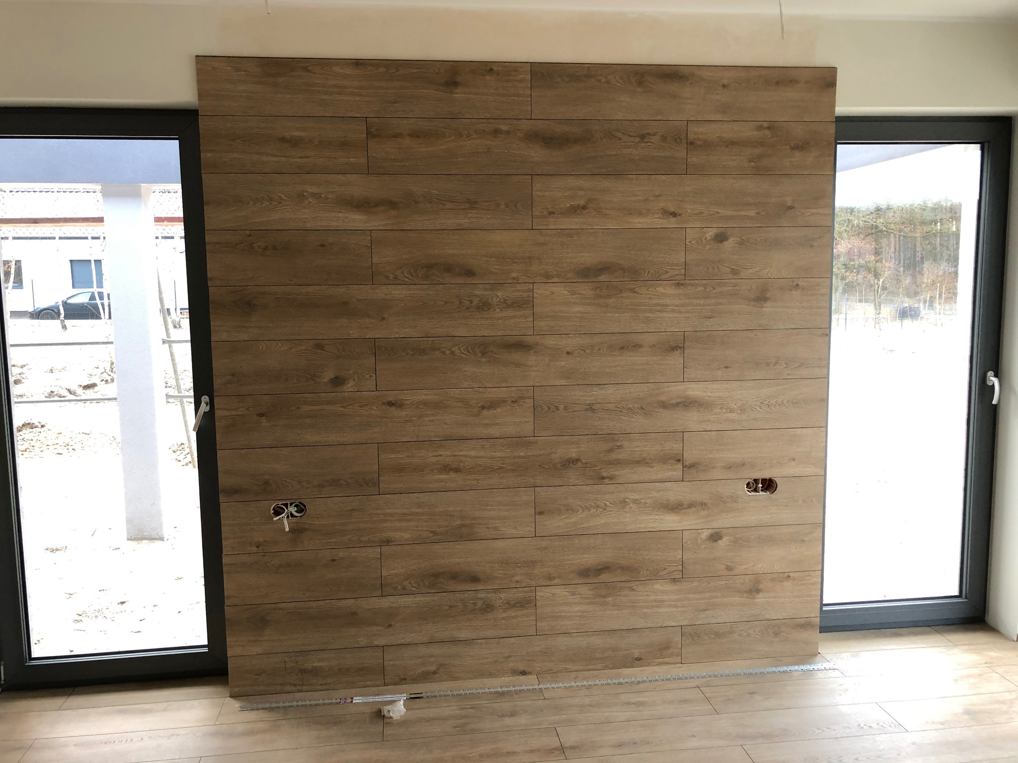 Ściana z paneli drewnopodobnych w jasnym wnętrzu, z widocznymi gniazdkami elektrycznymi i oknami po bokach. Podłoga również wykończona panelami.