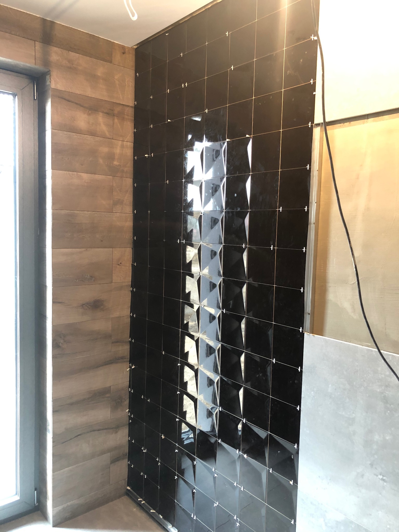 Częściowo wykończona ściana w łazience z błyszczącymi, czarnymi płytkami 3D i drewnopodobnymi panelami. Widoczne krzyżyki dystansowe.