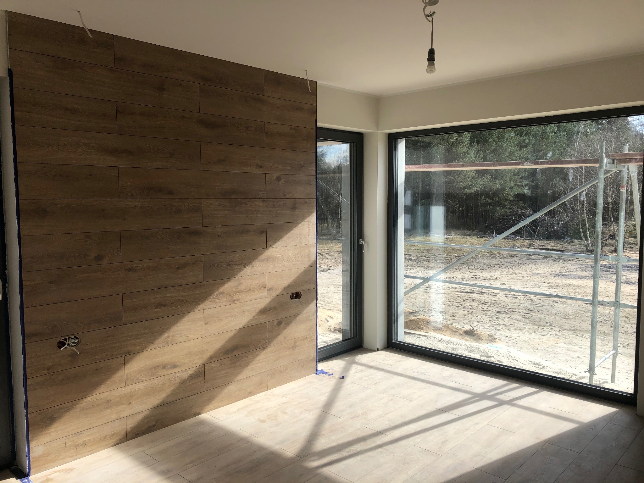 Wykończenie ściany panelami drewnopodobnymi w jasnym wnętrzu z dużym oknem. Widoczne gniazdko elektryczne i fragment podłogi z paneli. Na zewnątrz rusztowanie.