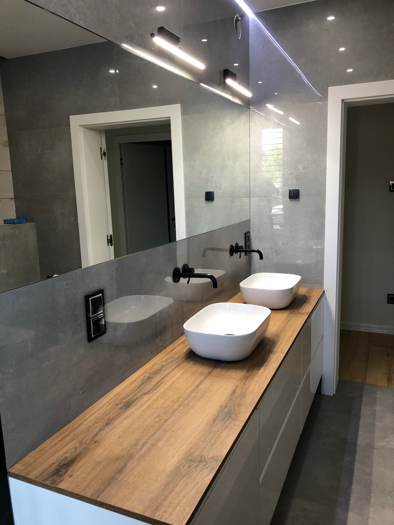 Nowoczesna łazienka z drewnianym blatem, dwiema białymi umywalkami i czarną armaturą na tle szarej ściany z lustrem i oświetleniem punktowym.