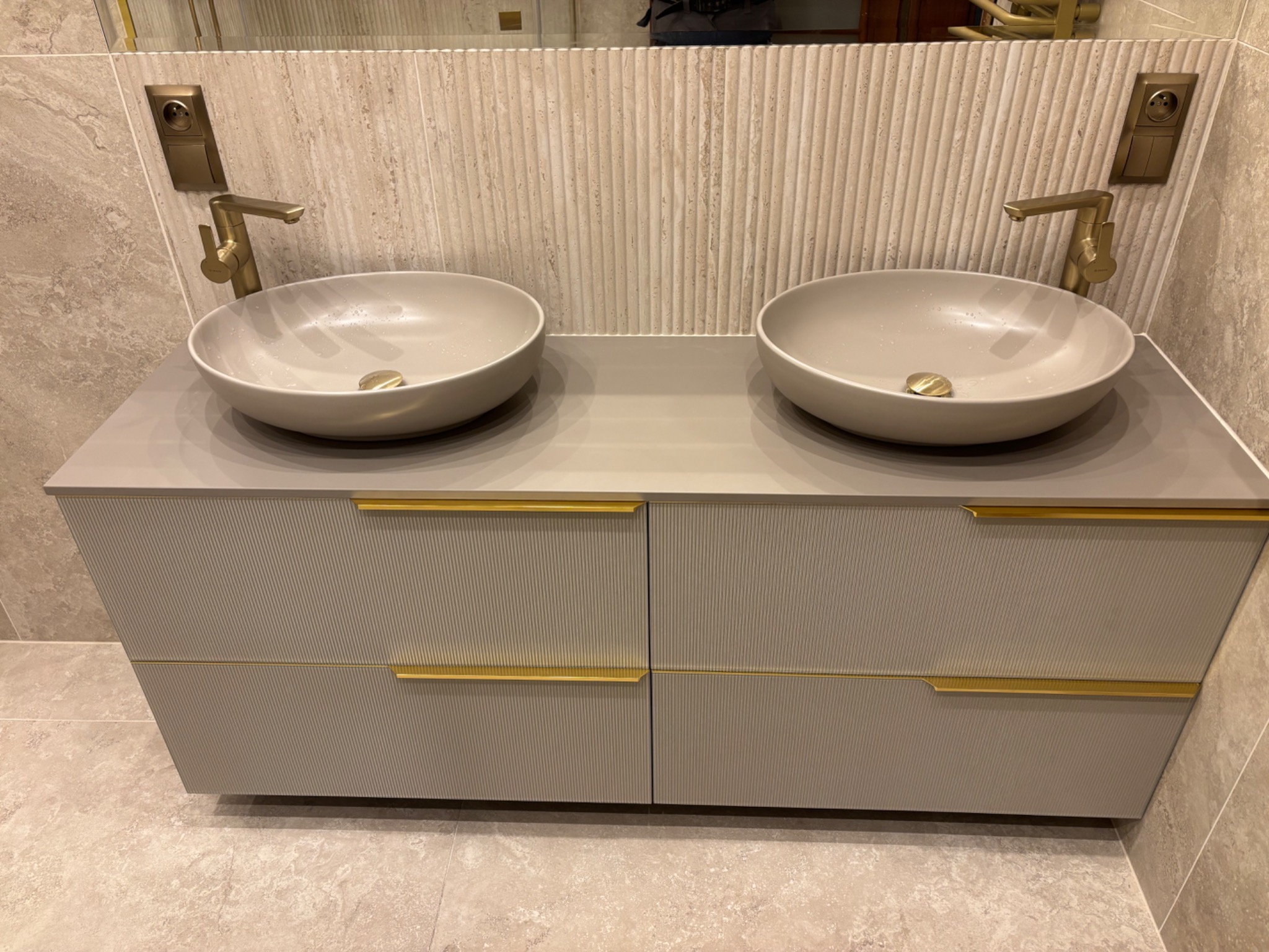 Elegancka łazienka: dwie umywalki nablatowe w odcieniach beżu z złotymi bateriami, szafka z ryflowanym frontem i złotymi uchwytami, płytki imitujące trawertyn.
