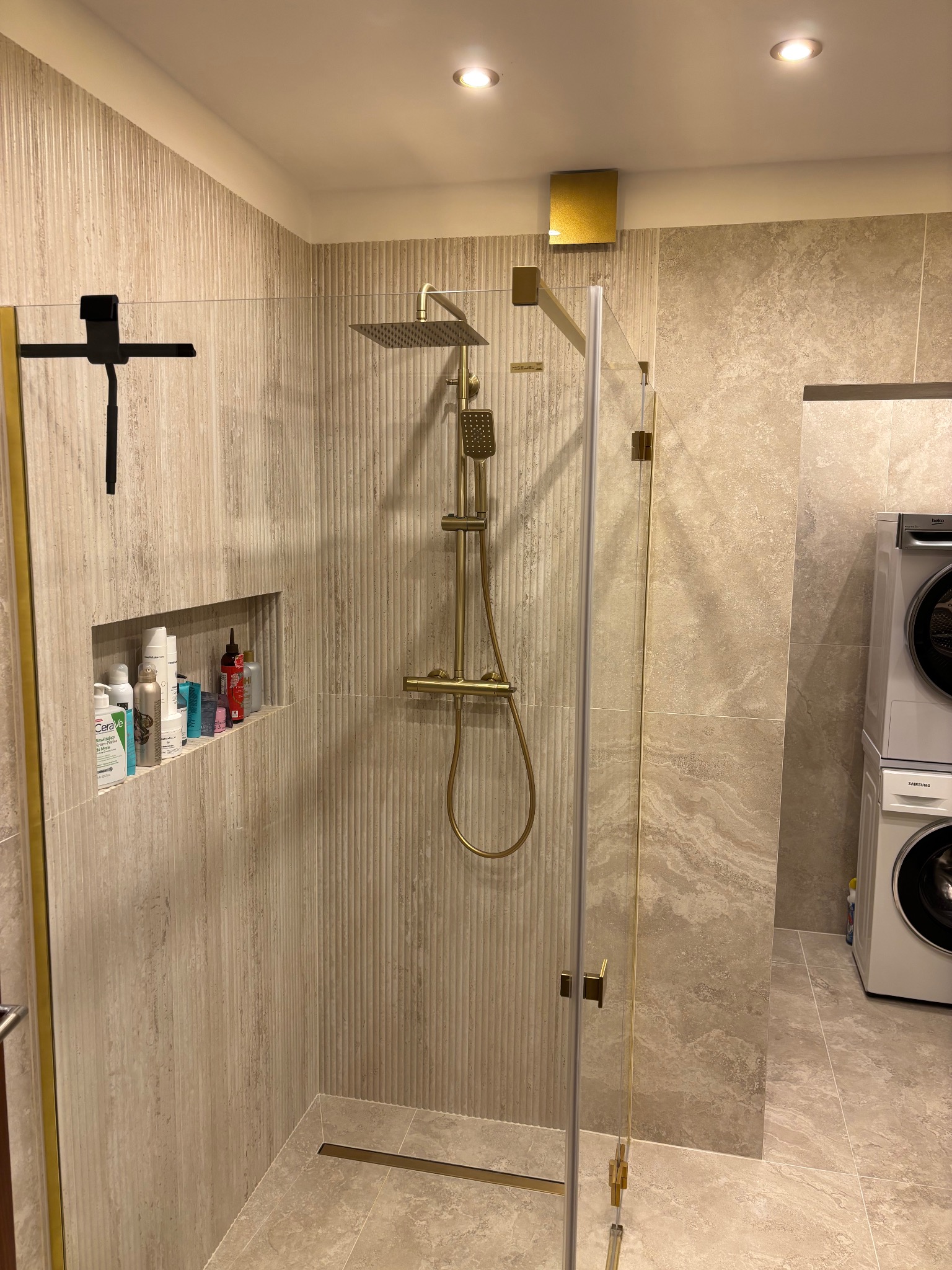 Beżowa łazienka z kabiną prysznicową typu walk-in, złotą armaturą i wnęką na kosmetyki. W tle pralka i suszarka.
