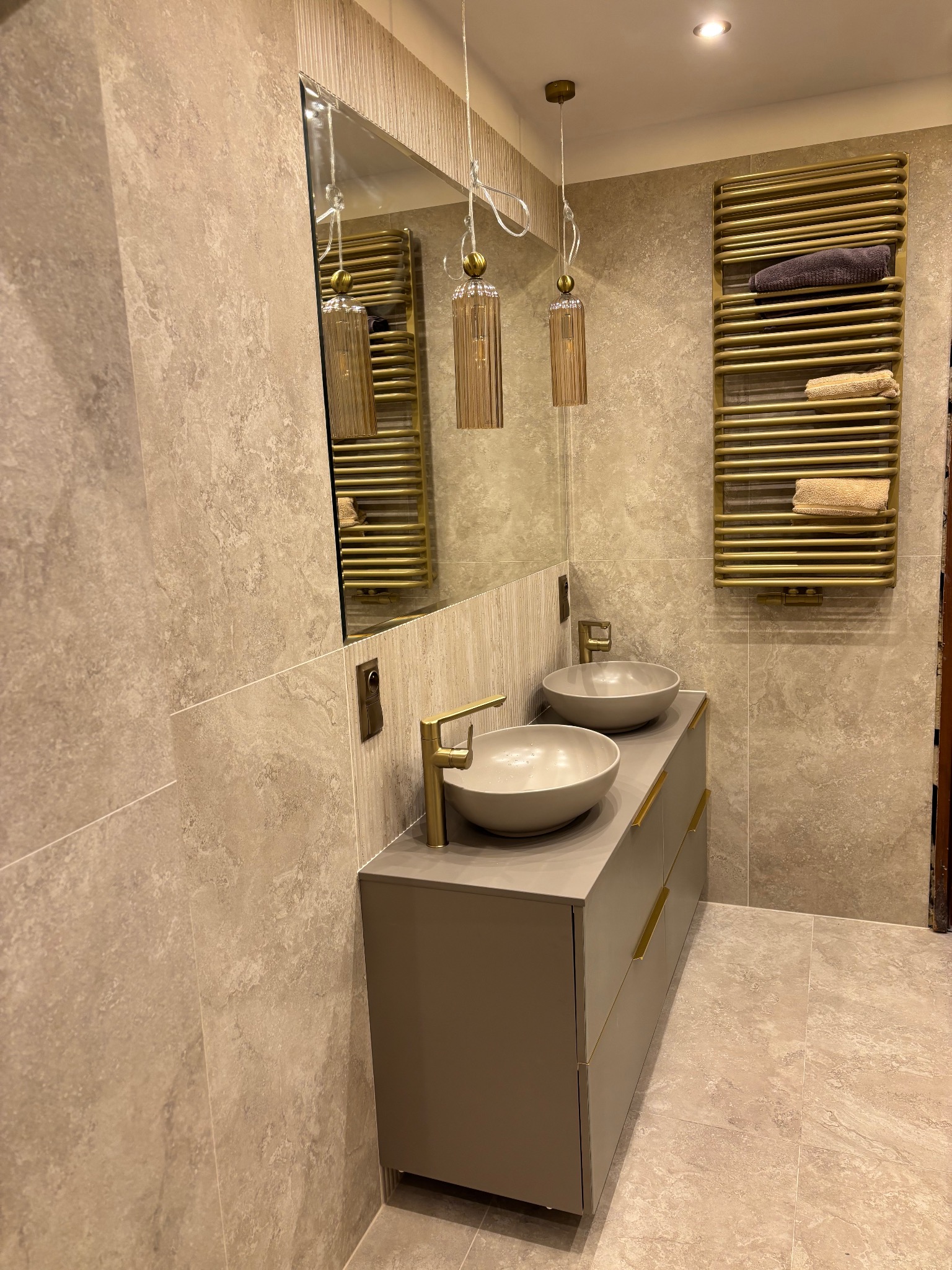 Nowoczesna łazienka z umywalkami nablatowymi, złotą armaturą i grzejnikiem drabinkowym. Beżowe płytki imitujące kamień na ścianach i podłodze. Lustro i wiszące lampy.