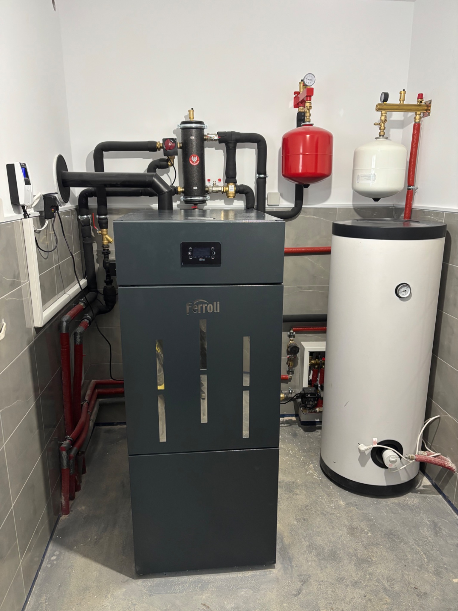 Nowoczesny piec CO marki Ferroli z instalacją grzewczą, zbiornikiem i czarnymi rurami w kotłowni wykończonej szarą płytką. Widoczne elementy instalacji i manometry.