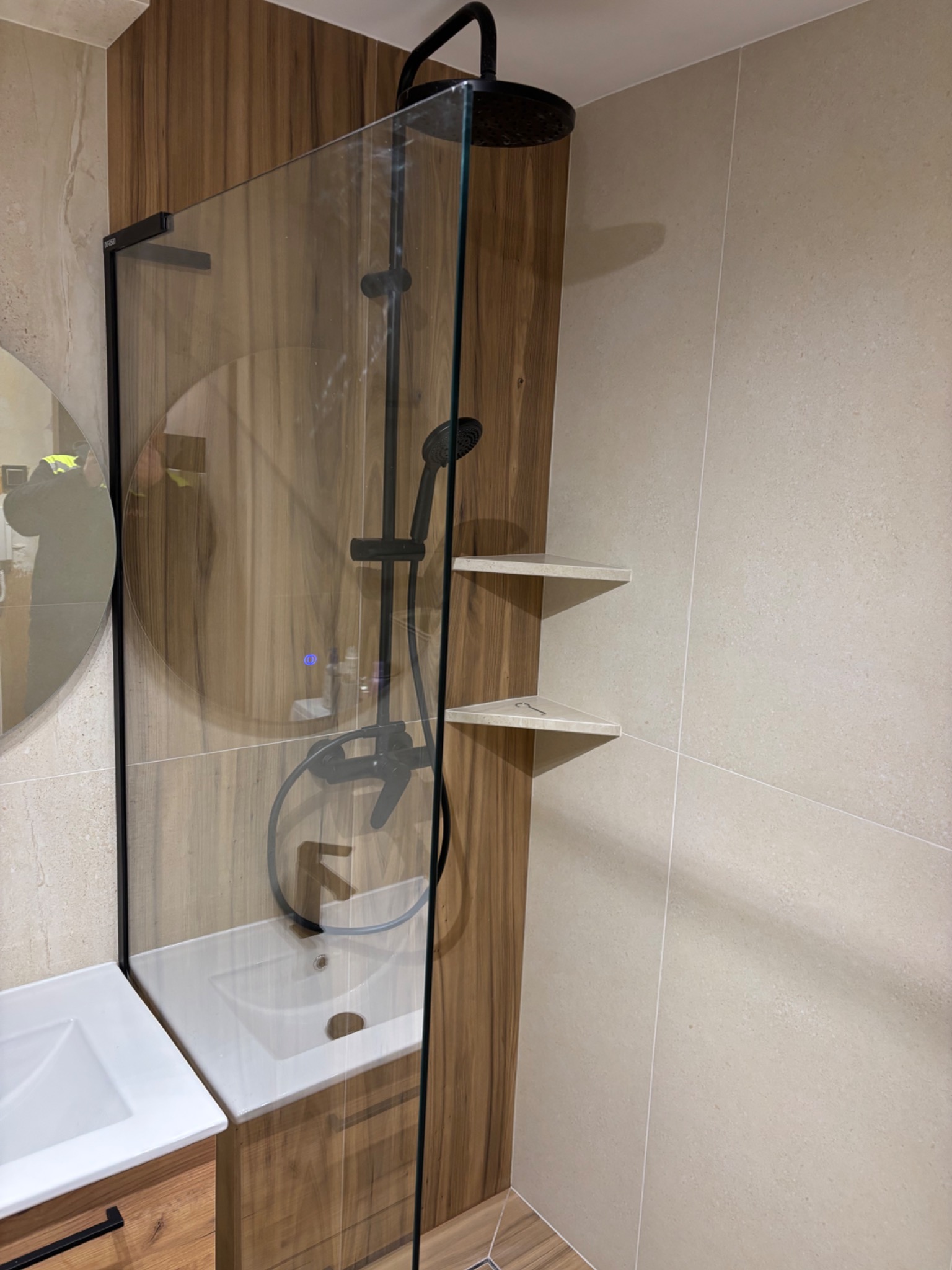 Nowoczesna kabina prysznicowa typu walk-in z czarną armaturą i drewnopodobnymi panelami ściennymi, uzupełniona o półki. Widoczna umywalka z szafką.