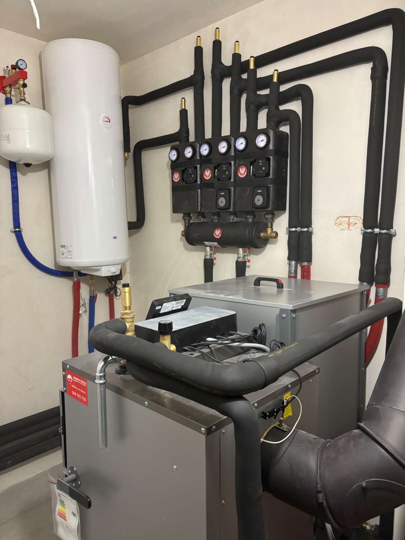 Złożona instalacja grzewcza w kotłowni: zasobnik ciepłej wody, rozdzielacz z manometrami, kocioł z czarnymi rurami izolowanymi termicznie. Realizacja instalacji grzewczej.