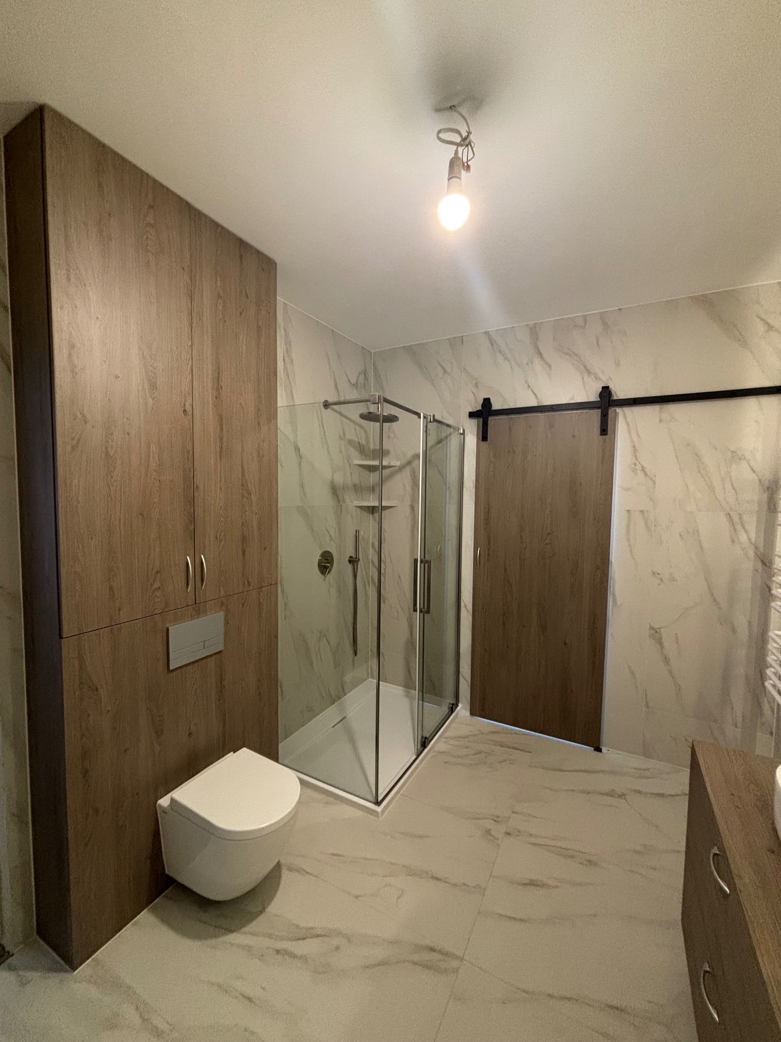 Nowoczesna łazienka z kabiną prysznicową, podwieszaną toaletą i przesuwnymi drzwiami w stylu stodoły. Marmurowe płytki na ścianach i podłodze, drewniane szafki.