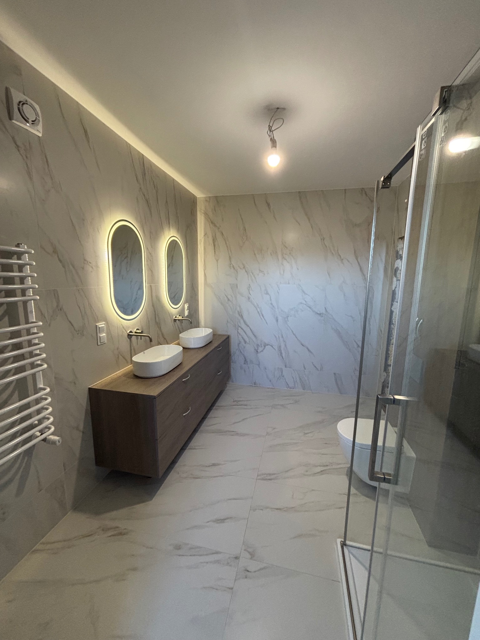 Nowoczesna łazienka z marmurowymi płytkami, podświetlanymi lustrami owalnymi i drewnianą szafką pod umywalki. Widoczna kabina prysznicowa i grzejnik drabinkowy.