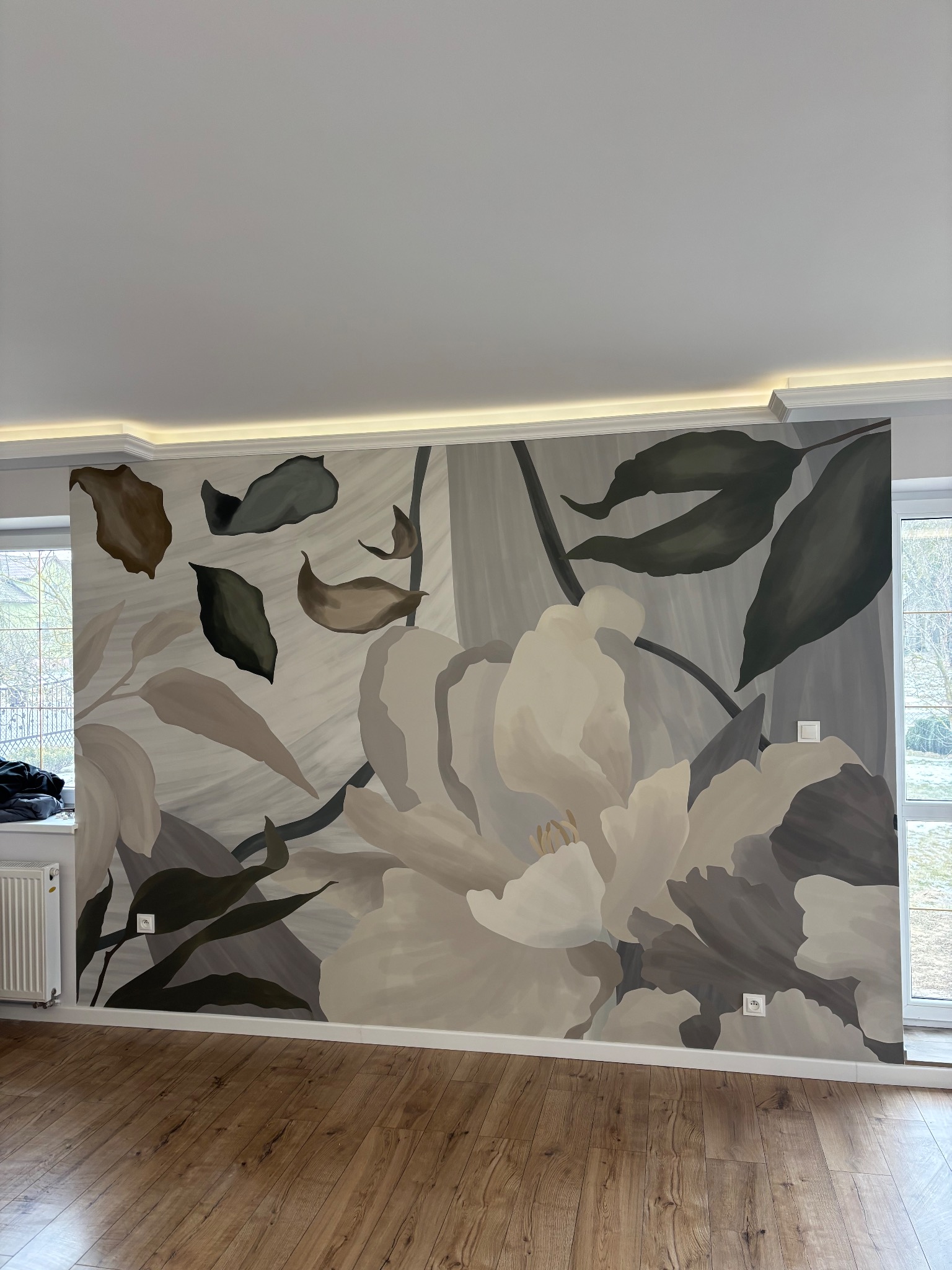 Ściana z ręcznie malowanym, abstrakcyjnym motywem kwiatowym w odcieniach szarości i beżu, drewniana podłoga, okno z widokiem na zieleń. Dekoracyjne oświetlenie sufitowe.