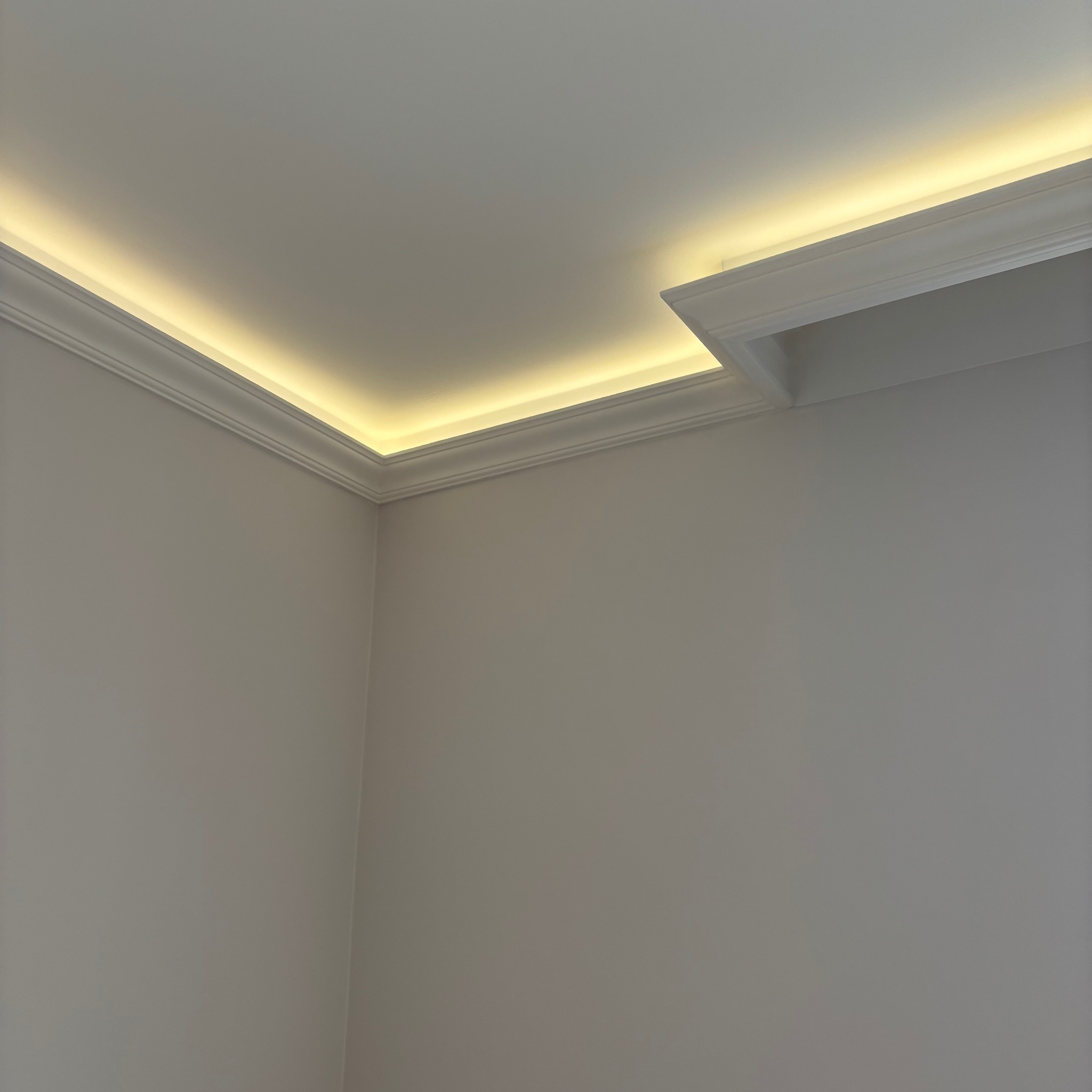 Nowoczesne oświetlenie LED wzdłuż sufitu z dekoracyjną listwą, tworzące ciepłą, subtelną poświatę na gładkich, jasnych ścianach. Minimalistyczny design.