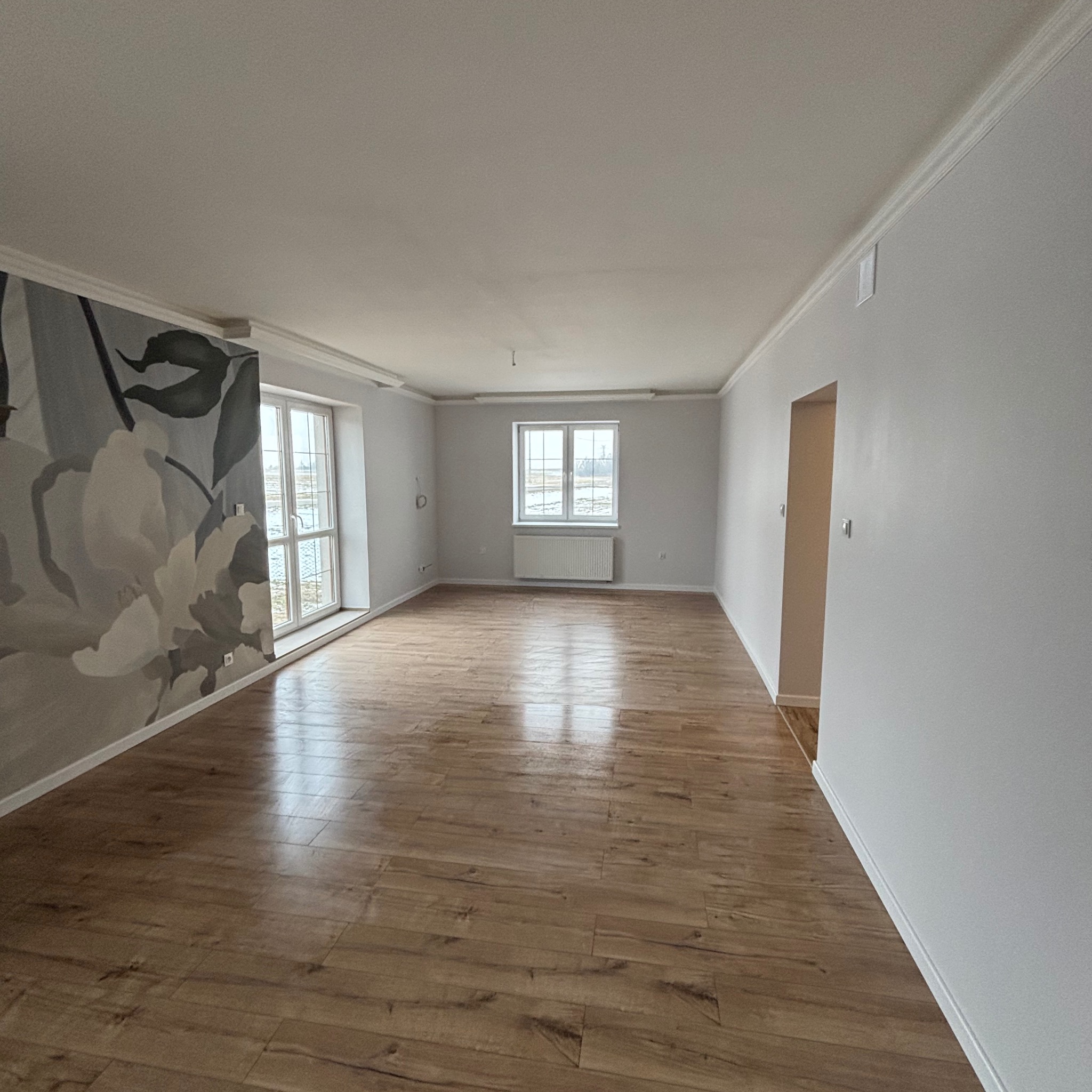 Jasne wnętrze po remoncie: drewniana podłoga, białe ściany z dekoracyjnym muralem kwiatowym, okna z widokiem na zewnątrz. Minimalistyczny design, świeżo po wykończeniu.