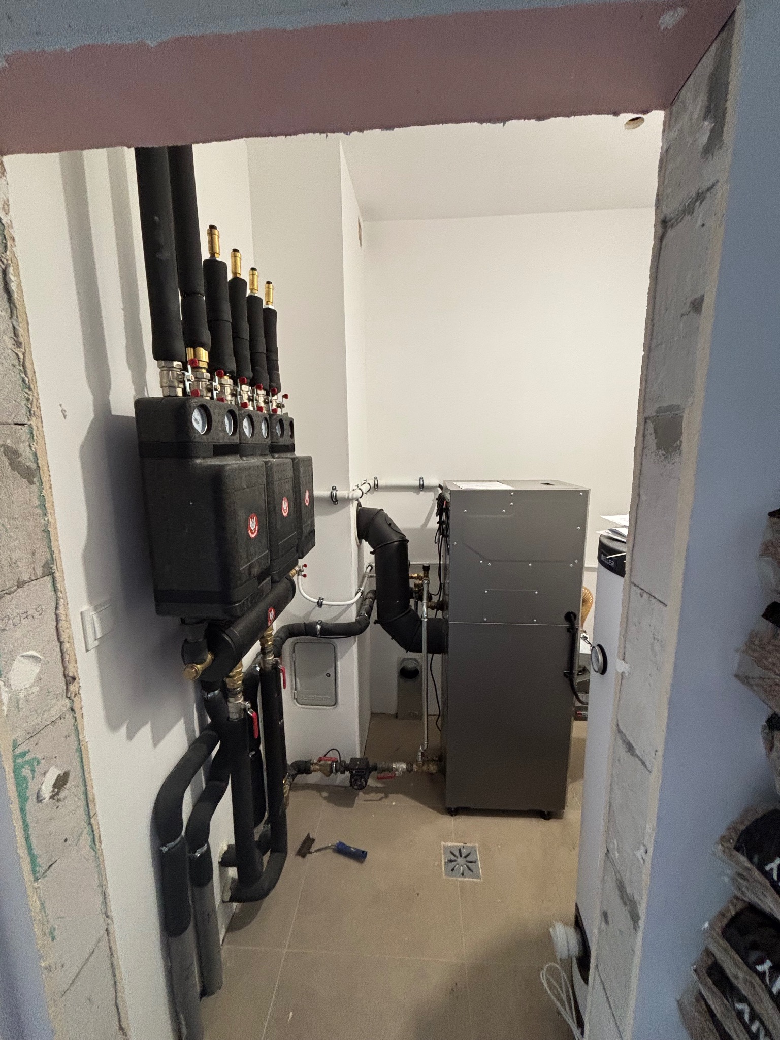 Zainstalowany system grzewczy: czarny rozdzielacz z manometrami i izolowanymi rurami, obok szare urządzenie grzewcze na płytkach w pomieszczeniu technicznym.