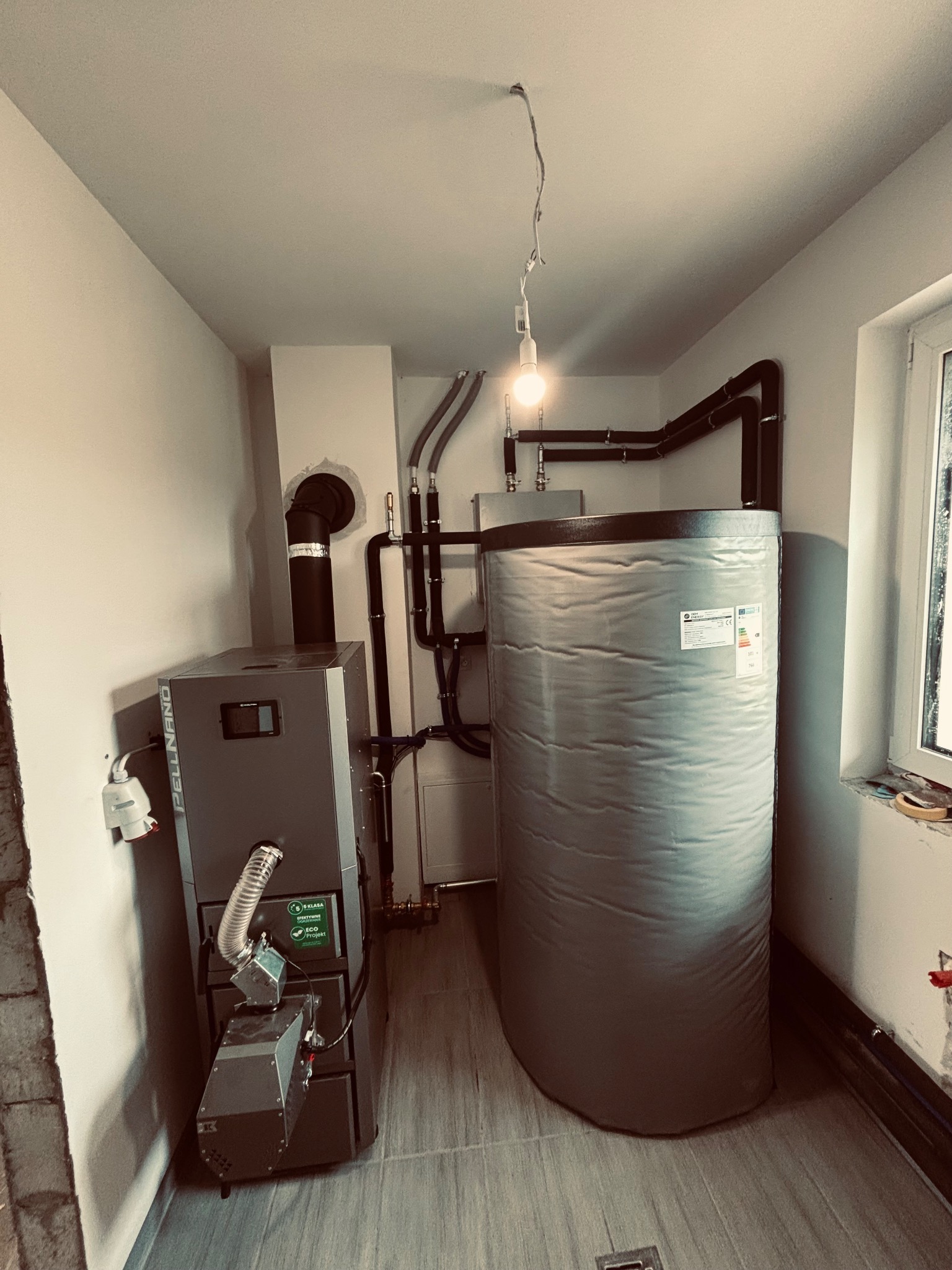 Nowoczesna kotłownia z szarym piecem pelletowym Pellnano i dużym zasobnikiem ciepłej wody z etykietą energetyczną. Estetycznie poprowadzone czarne rury w jasnym pomieszczeniu z oknem, tymczasowym...