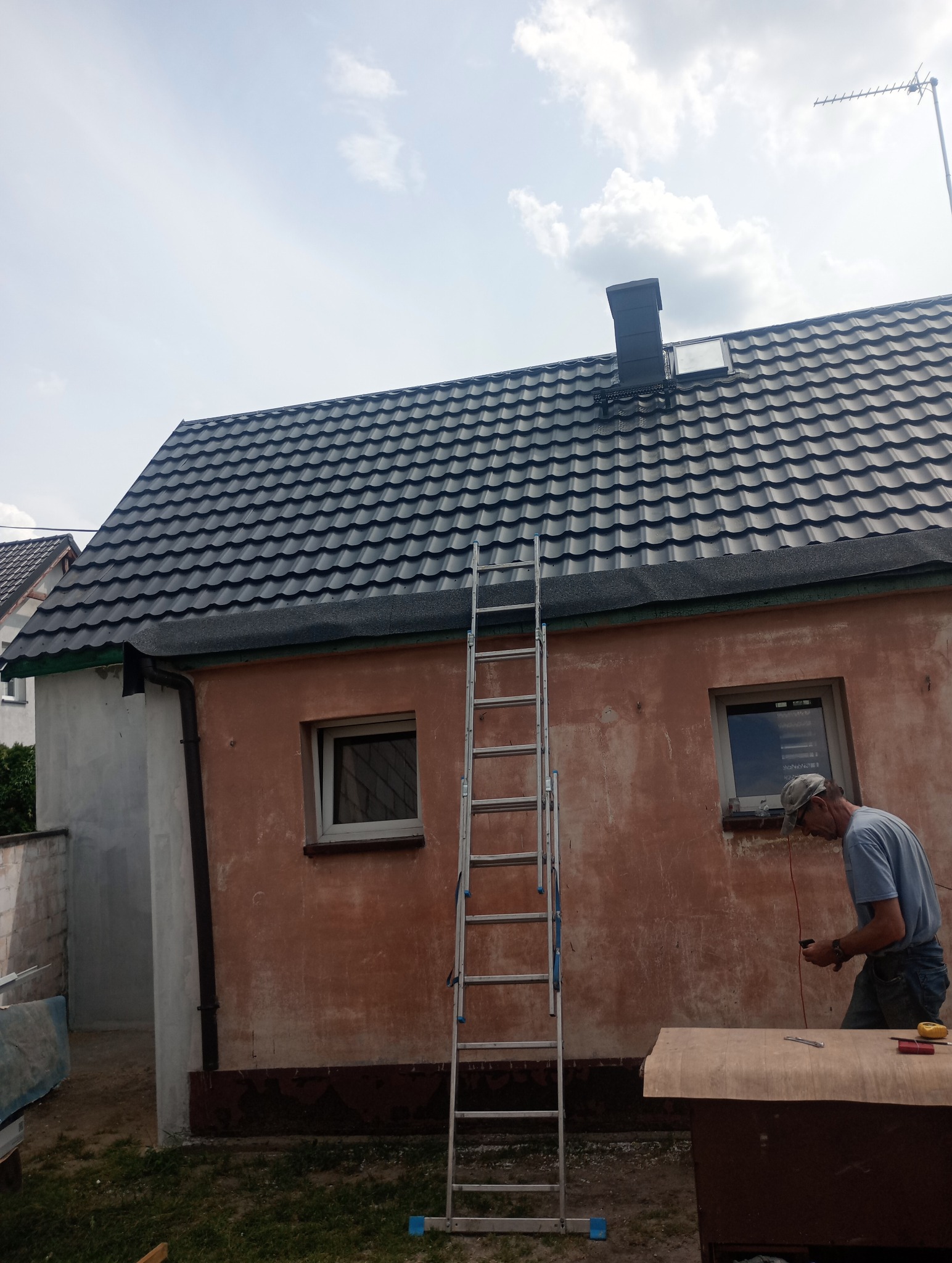 Praca przy dachu: dekarz przygotowuje materiały na tle domu z nowym, ciemnym dachem, widoczna drabina oparta o ścianę i komin na dachu.