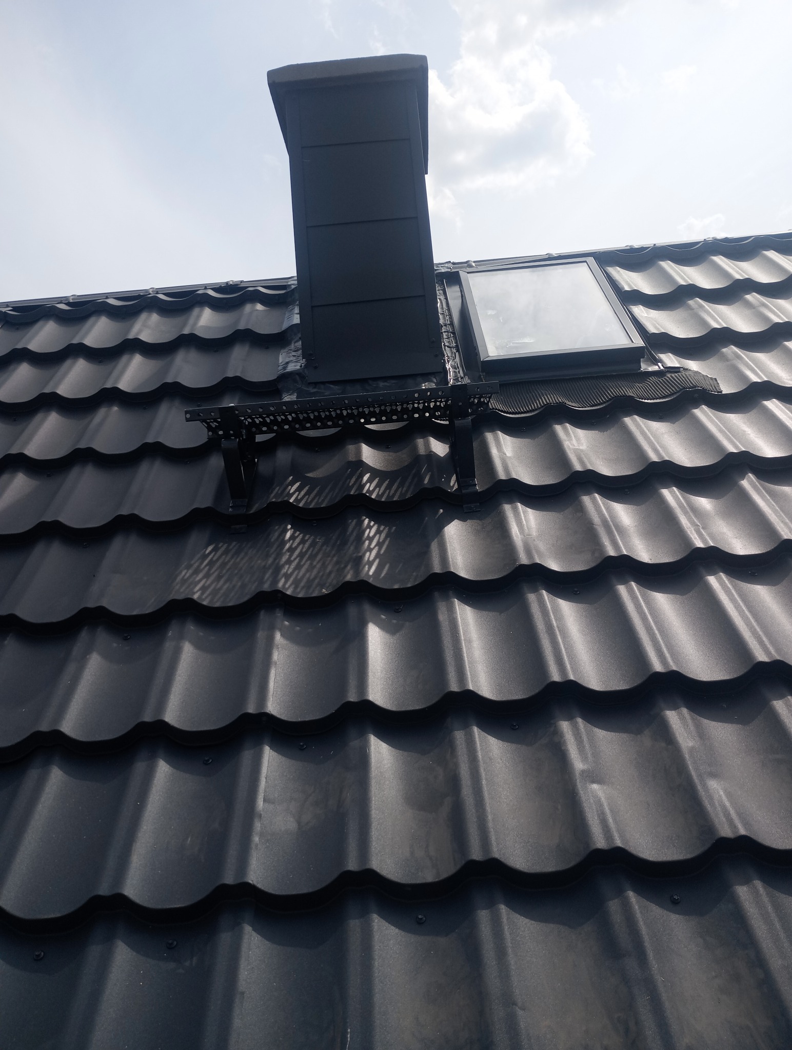 Czarny dach z blachy trapezowej z zamontowanym oknem dachowym i kominem wentylacyjnym, widoczne elementy obróbki blacharskiej i mocowania.