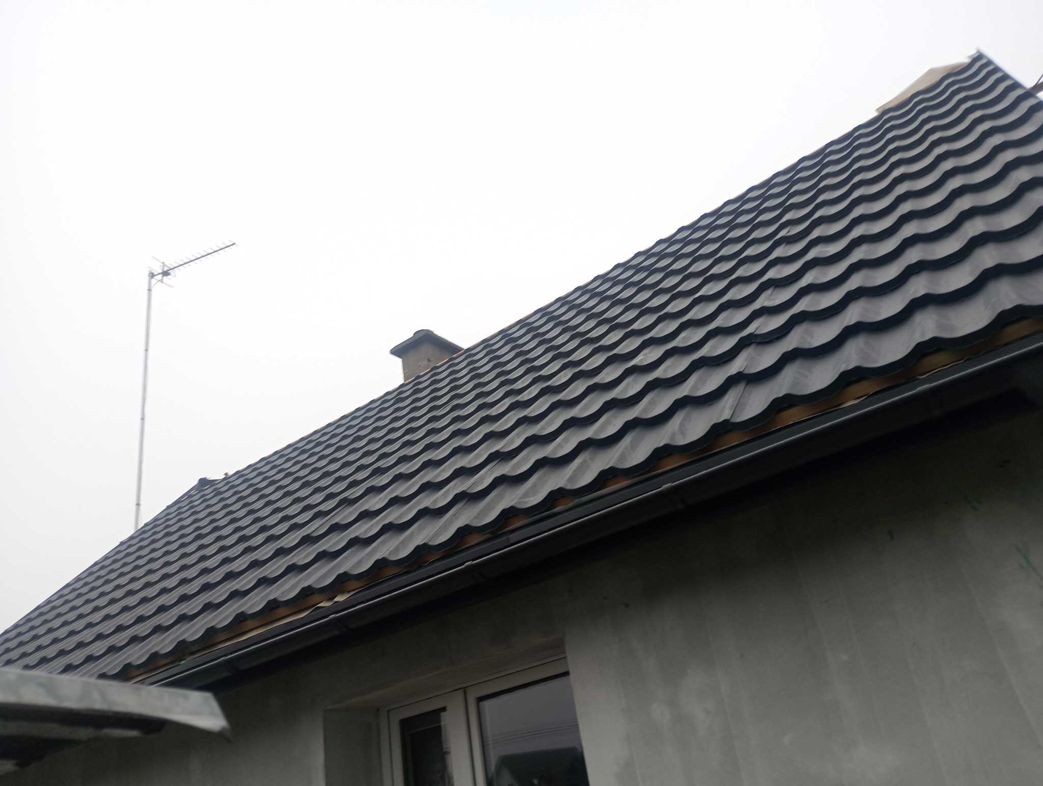 Nowy, grafitowy dach z blachodachówki modułowej, widoczny komin i antena telewizyjna, szara elewacja budynku z oknem.