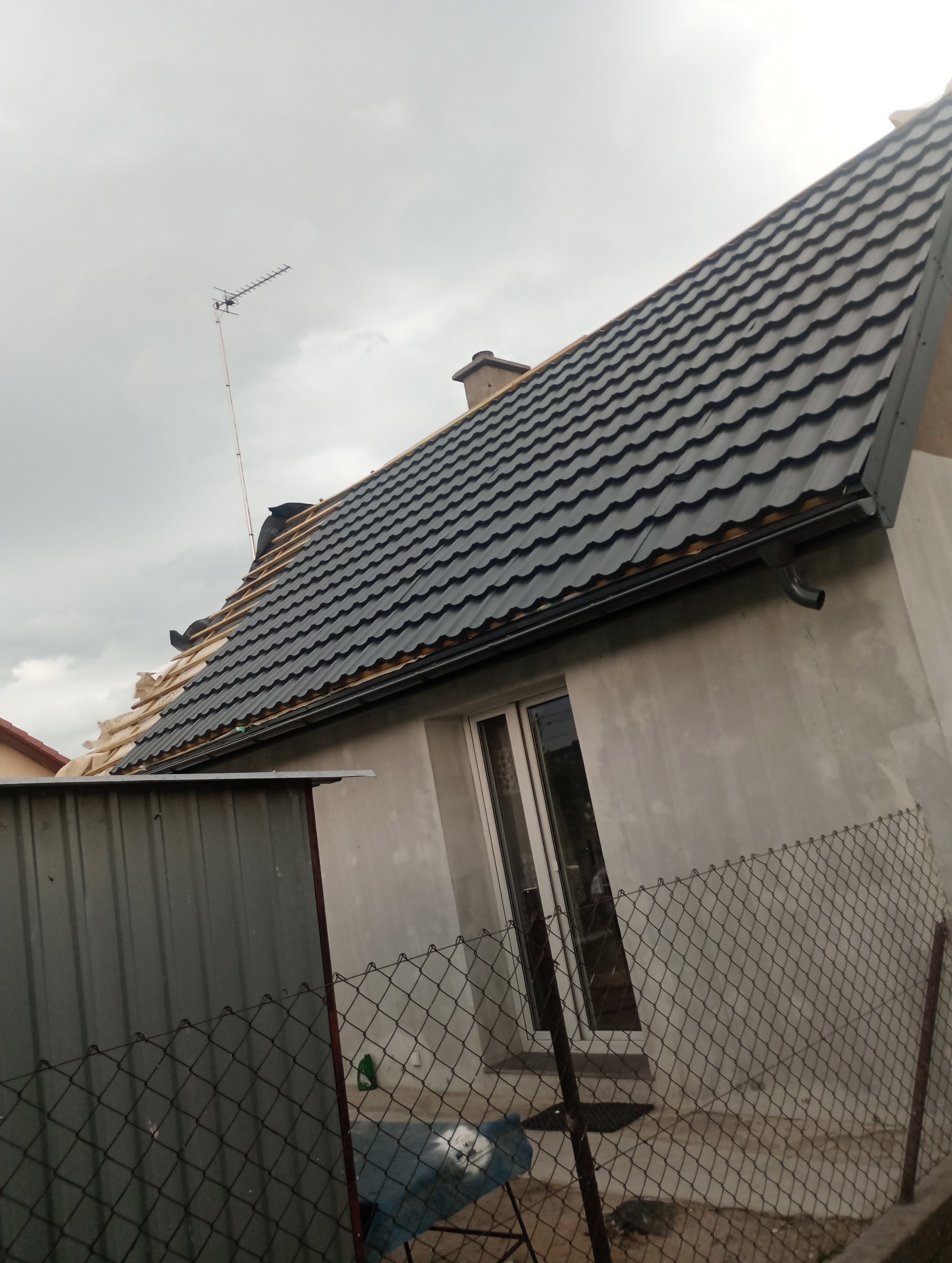 Częściowo pokryty dach z blachodachówki w kolorze grafitowym, widoczne elementy konstrukcji dachu przed ukończeniem, antena telewizyjna na tle pochmurnego nieba.