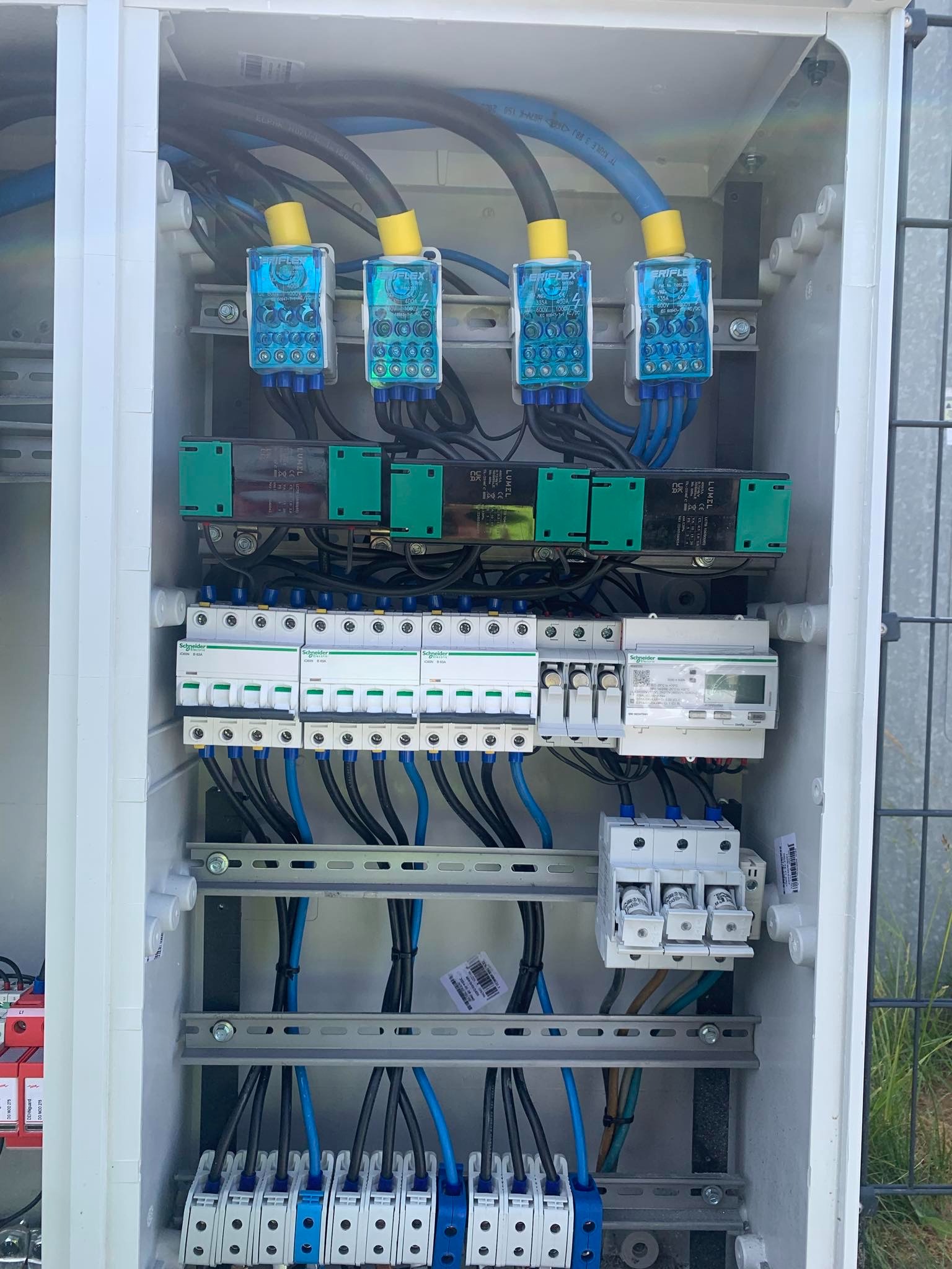 Wnętrze białej skrzynki elektrycznej z zamontowanymi bezpiecznikami, przewodami w kolorze czarnym i niebieskim, oraz elementami pomiarowymi. Widoczne złącza i listwy zaciskowe.