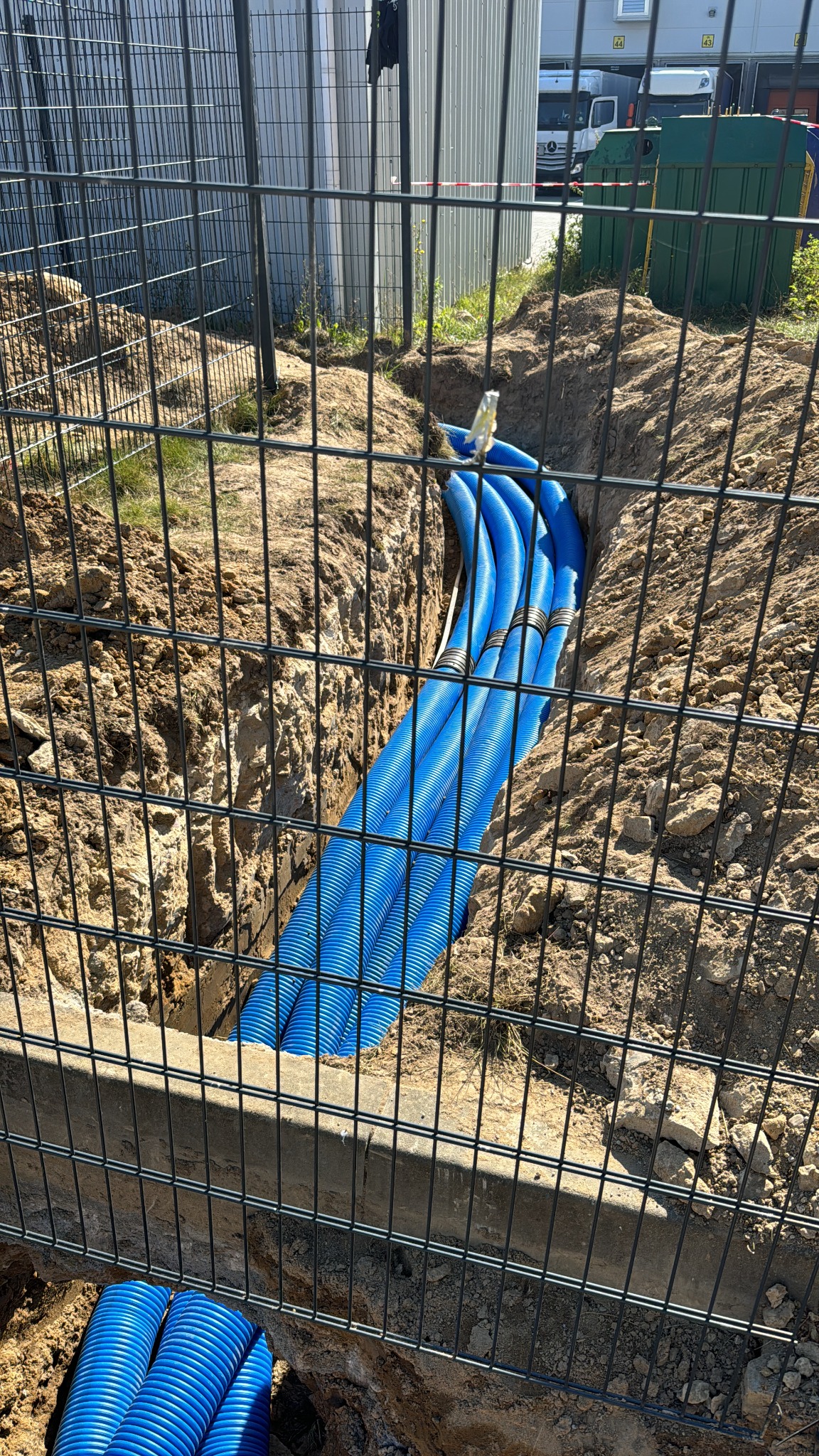 Błękitne rury karbowane ułożone w wykopie ziemnym, zabezpieczone siatką ogrodzeniową, widok z góry na instalację podziemną.