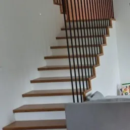 Nowoczesne schody z drewnianymi stopniami i czarną balustradą, widok z dołu. Minimalistyczny design, jasne wnętrze, fragment szarej sofy w kadrze.