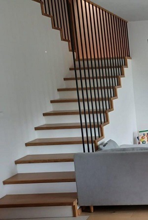 Nowoczesne schody z drewnianymi stopniami i czarną balustradą, widok z dołu. Minimalistyczny design, jasne wnętrze, fragment szarej sofy w kadrze.