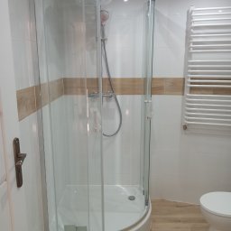 EdenBud - Nowa kabina prysznicowa narożna z chromowanymi profilami, zamontowana w wykończonej łazience z białymi płytkami i drewnopodobną podłogą, bateria prysznicowa w folii ochronnej.