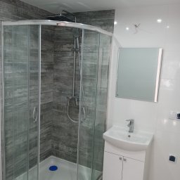 EdenBud - Nowoczesna łazienka po remoncie: kabina prysznicowa z szarymi panelami imitującymi drewno, biała umywalka z szafką i lustro na białych płytkach.