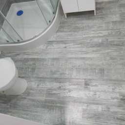 EdenBud - Wykończona łazienka z kabiną prysznicową, białą szafką i toaletą zabezpieczoną folią ochronną, podłoga wyłożona szarymi panelami imitującymi drewno.