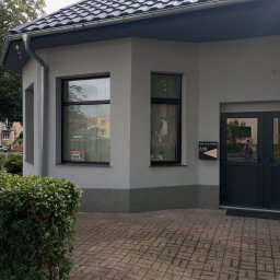 EdenBud - Wykończony budynek z szarą elewacją, ciemną stolarką okienną i nowym dachem z blachodachówki, widoczne tabliczki informacyjne na oknach i skrzynka pocztowa przy drzwiach.