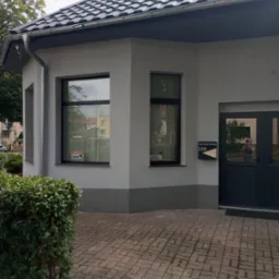 Wykończony budynek z szarą elewacją, ciemną stolarką okienną i nowym dachem z blachodach&oacute;wki, widoczne tabliczki informacyjne na oknach i skrzynka pocztowa przy drzwiach.