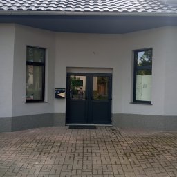 EdenBud - Elewacja budynku z szarym tynkiem, ciemnymi oknami i drzwiami wejściowymi, widoczna skrzynka pocztowa z numerem 21B.