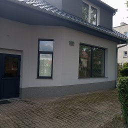 EdenBud - Szary budynek po remoncie elewacji, z ciemnymi oknami i drzwiami, widoczny z zewnątrz na tle kostki brukowej.