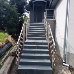 EdenBud - Zewnętrzne schody wejściowe wyłożone grafitową płytką z metalową barierką, prowadzące do ciemnoszarych drzwi wejściowych z zadaszeniem