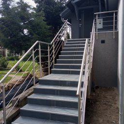 EdenBud - Zewnętrzne schody wejściowe wyłożone szarą płytką z metalowymi krawędziami i balustradą ze stali nierdzewnej prowadzące do szarego budynku z zadaszeniem.