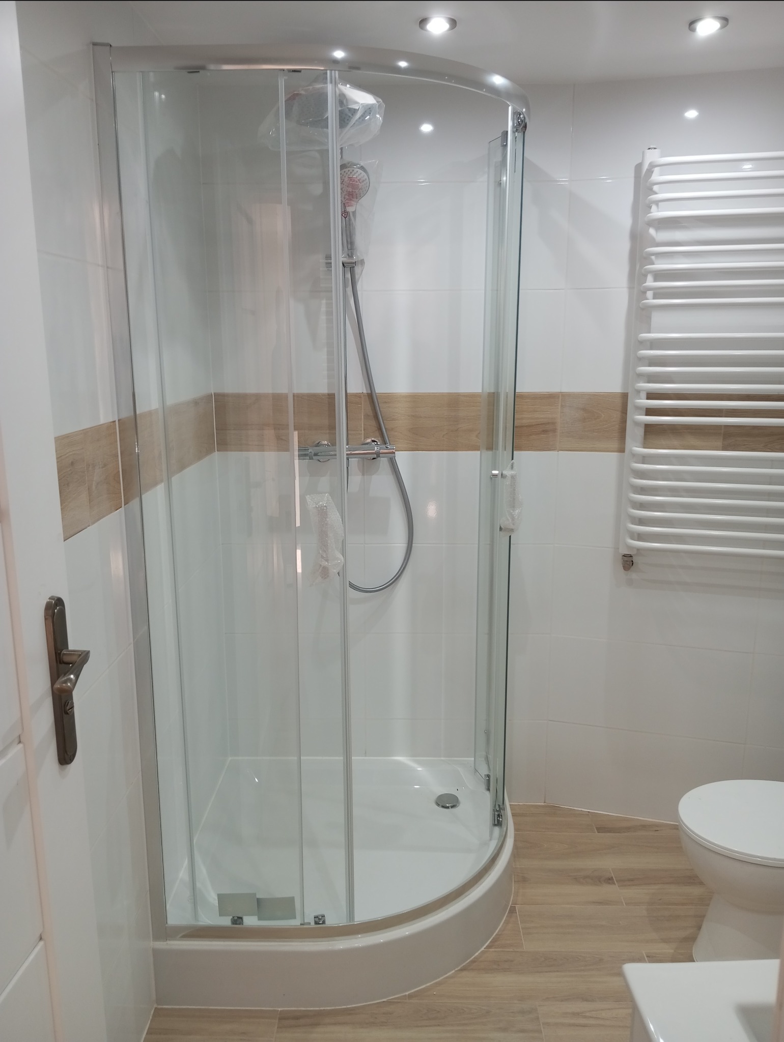 Nowa kabina prysznicowa narożna z chromowanymi profilami, zamontowana w wykończonej łazience z białymi płytkami i drewnopodobną podłogą, bateria prysznicowa w folii ochronnej.