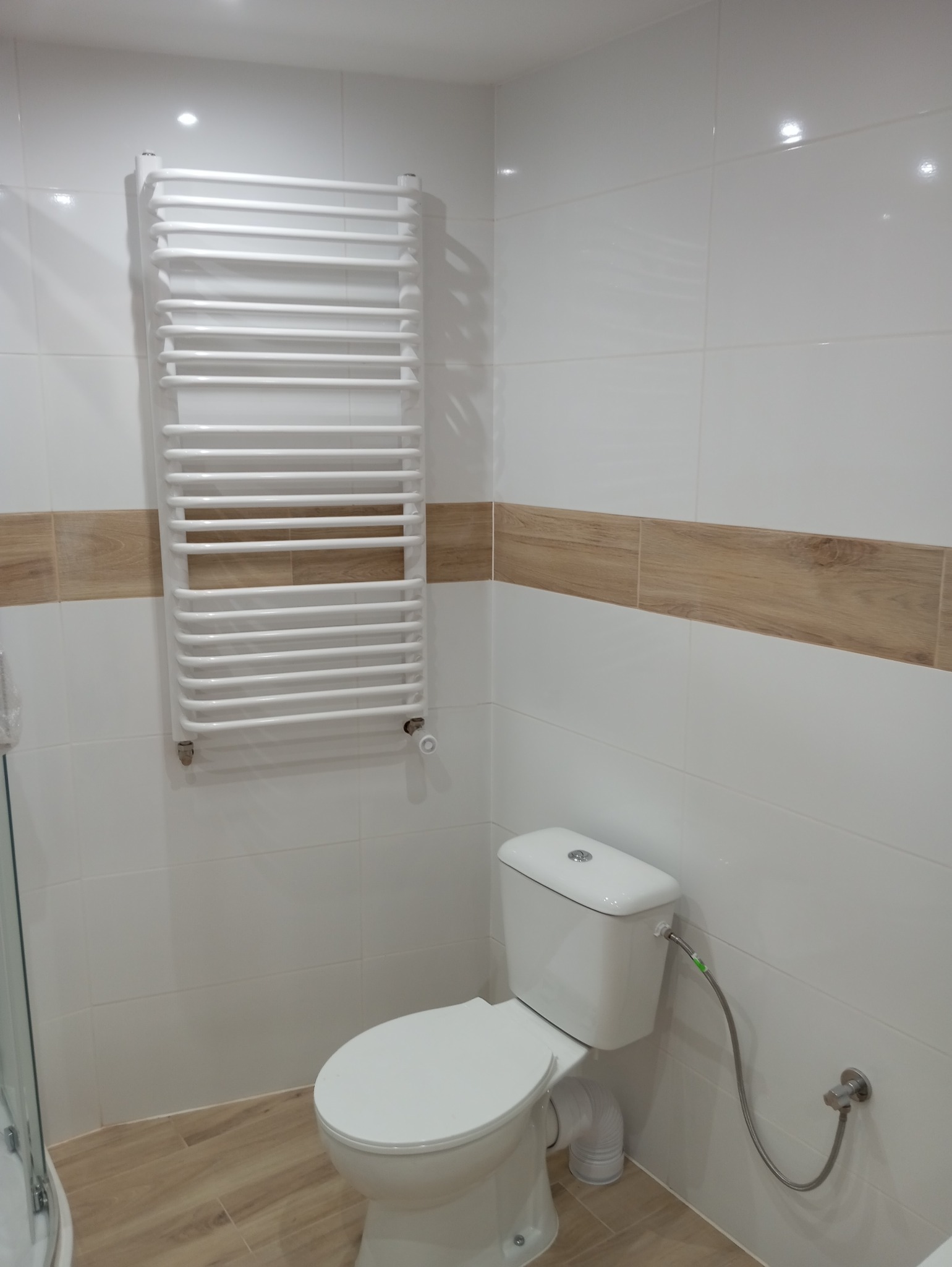 Biała łazienka z grzejnikiem drabinkowym, toaletą i dekoracyjnym pasem z płytek imitujących drewno.