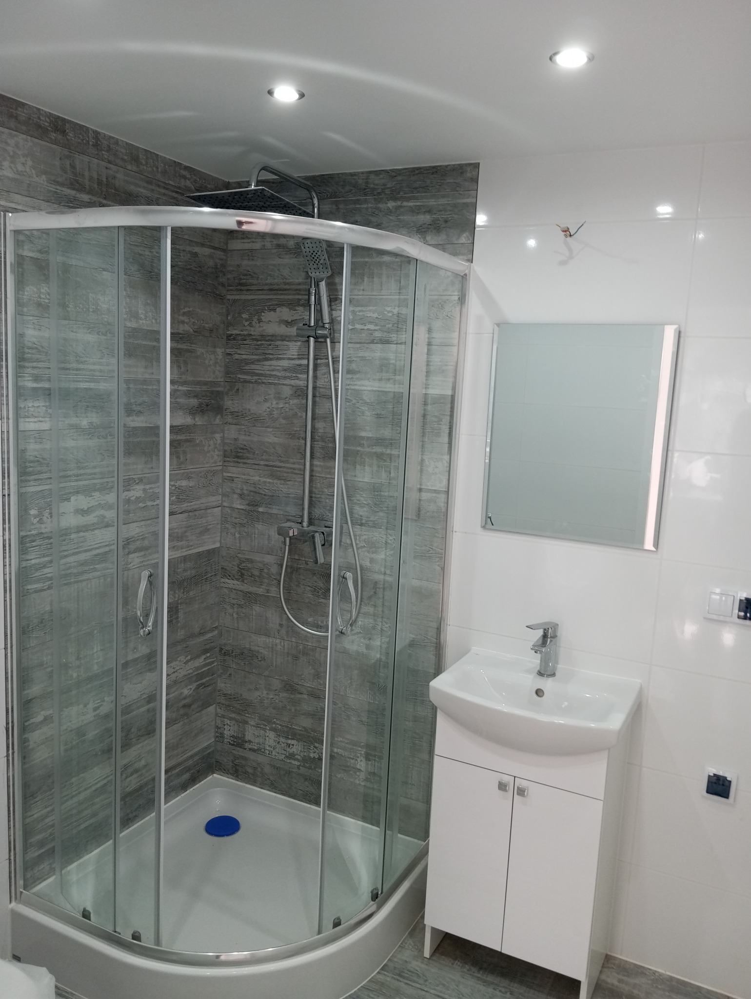 Nowoczesna łazienka po remoncie: kabina prysznicowa z szarymi panelami imitującymi drewno, biała umywalka z szafką i lustro na białych płytkach.